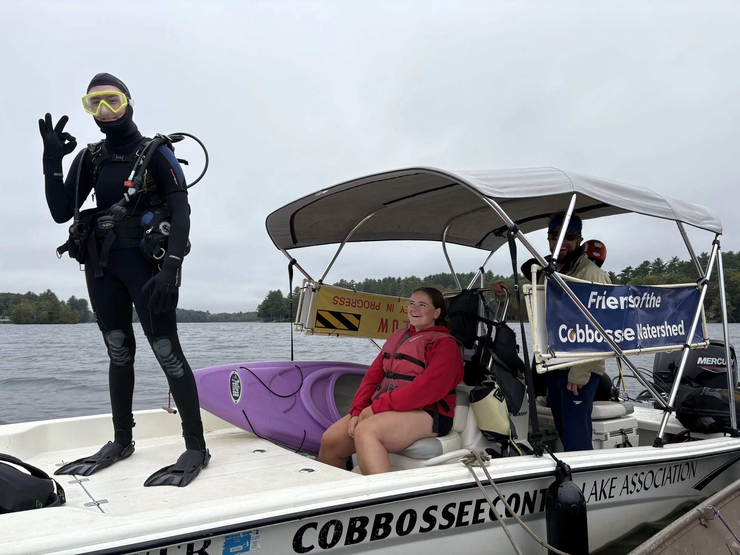 A person in full SCUBA gear standing on the deck of a boat giving the "okay" symbol, with a person in a red sweatshirt and life jacket smiling looking up at the diver and a purple kayak next to them