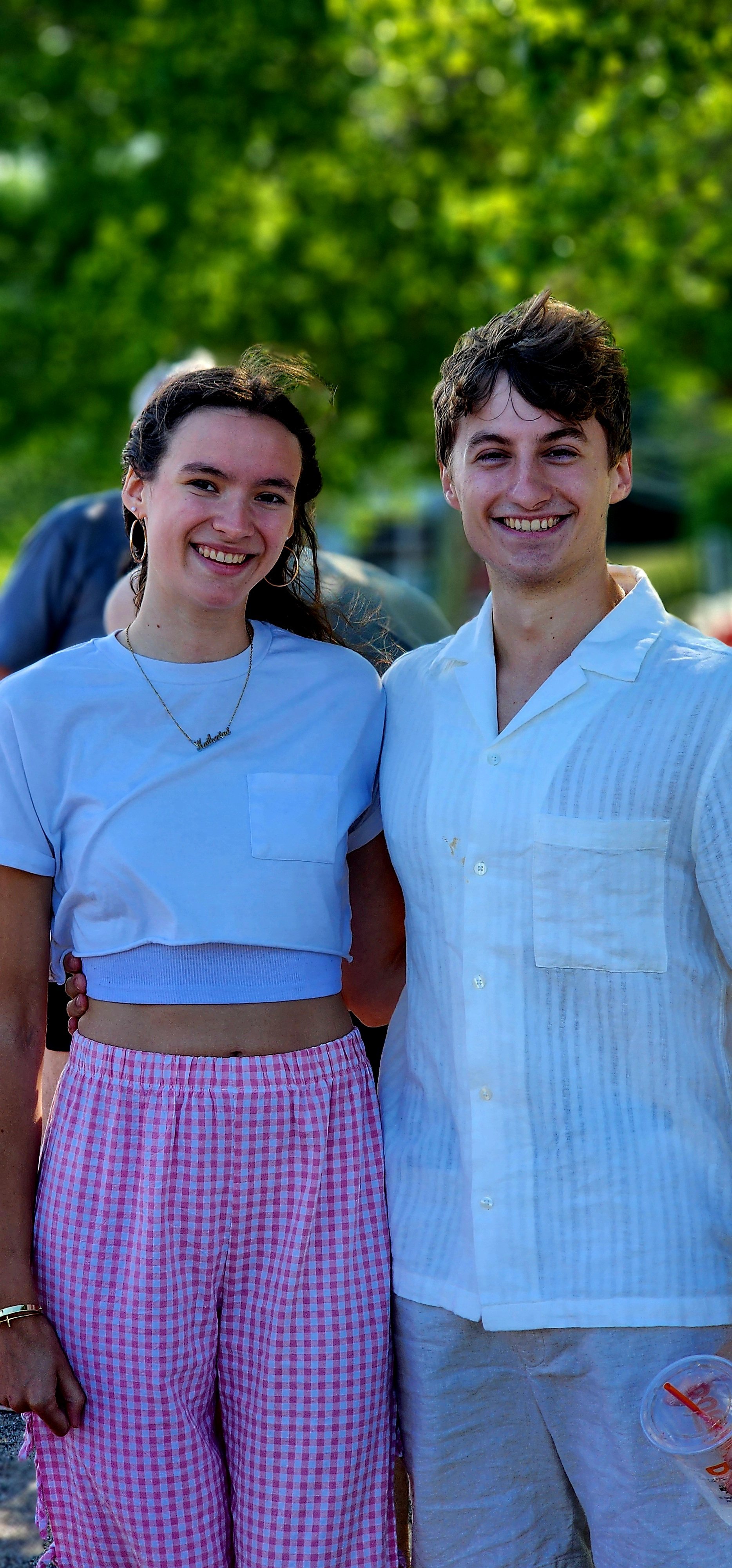  A woman wearing a white t-shirt and red gingham pants standing with a man wearing a white button up shirts and white shorts, both smiling at the camera