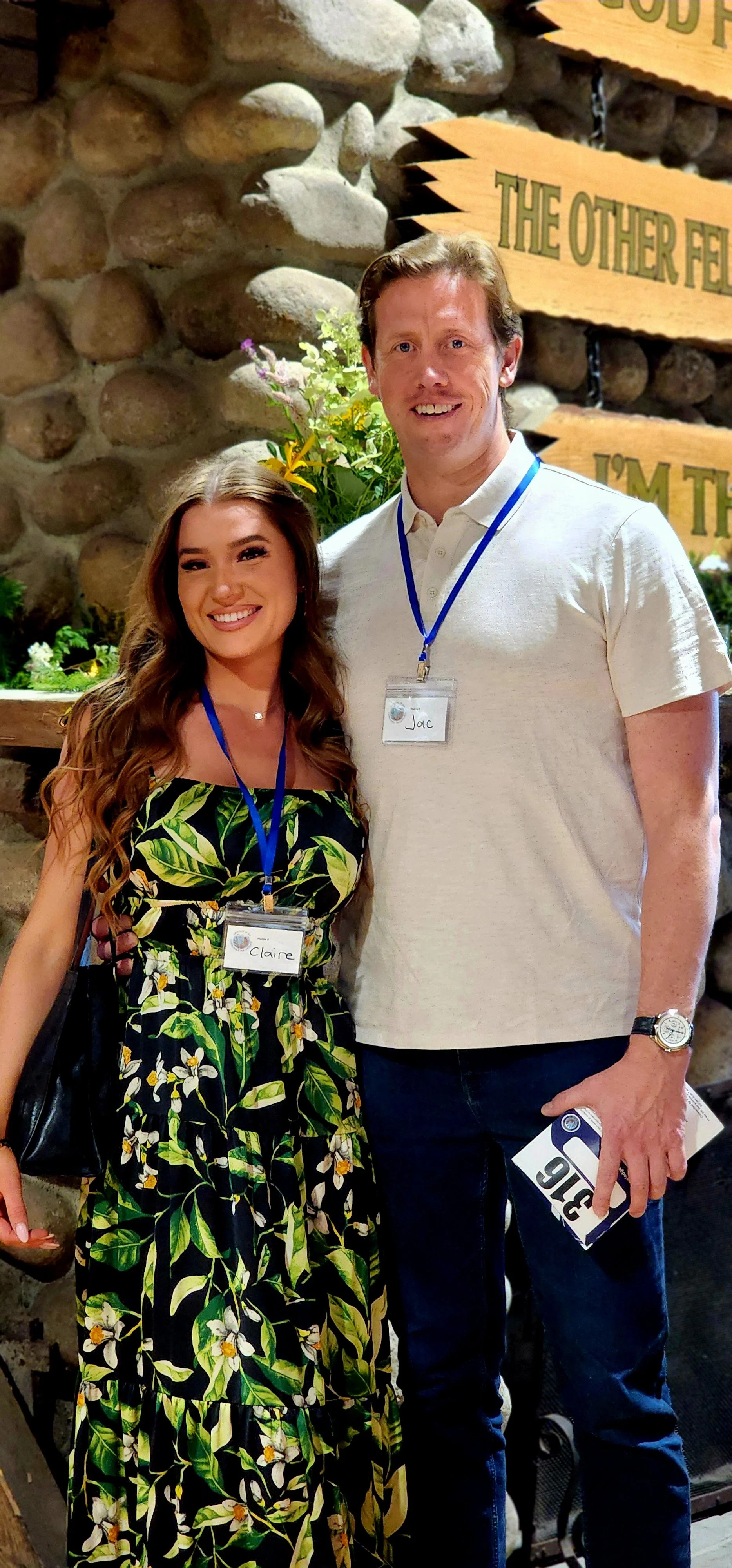 A woman, wearing a black dress with a green leaf pattern, and a man, wearing a white polo and jeans, smiling at the camera
