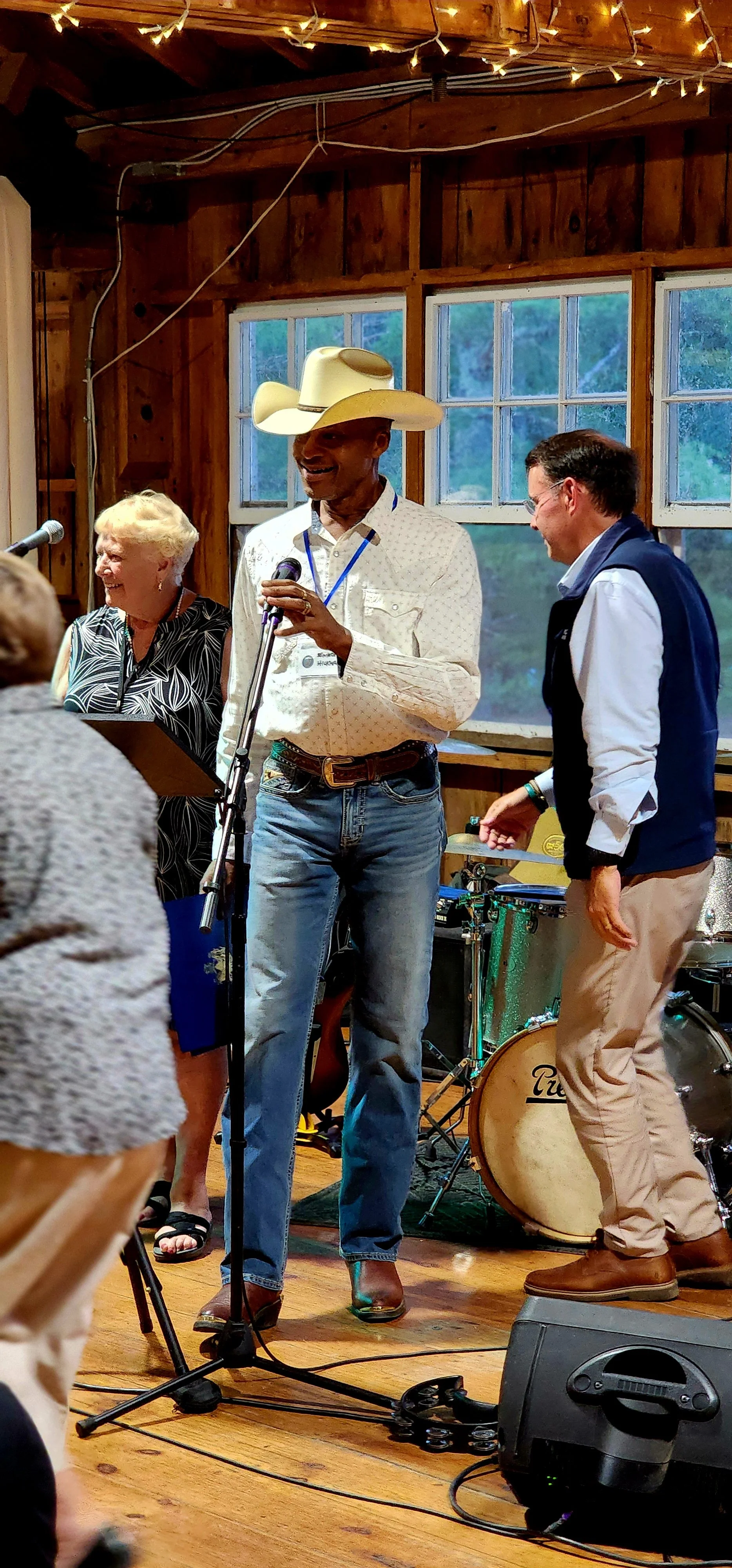 A man, wearing a white cowboy hat, white shirt and jeans, speaking into a microphone