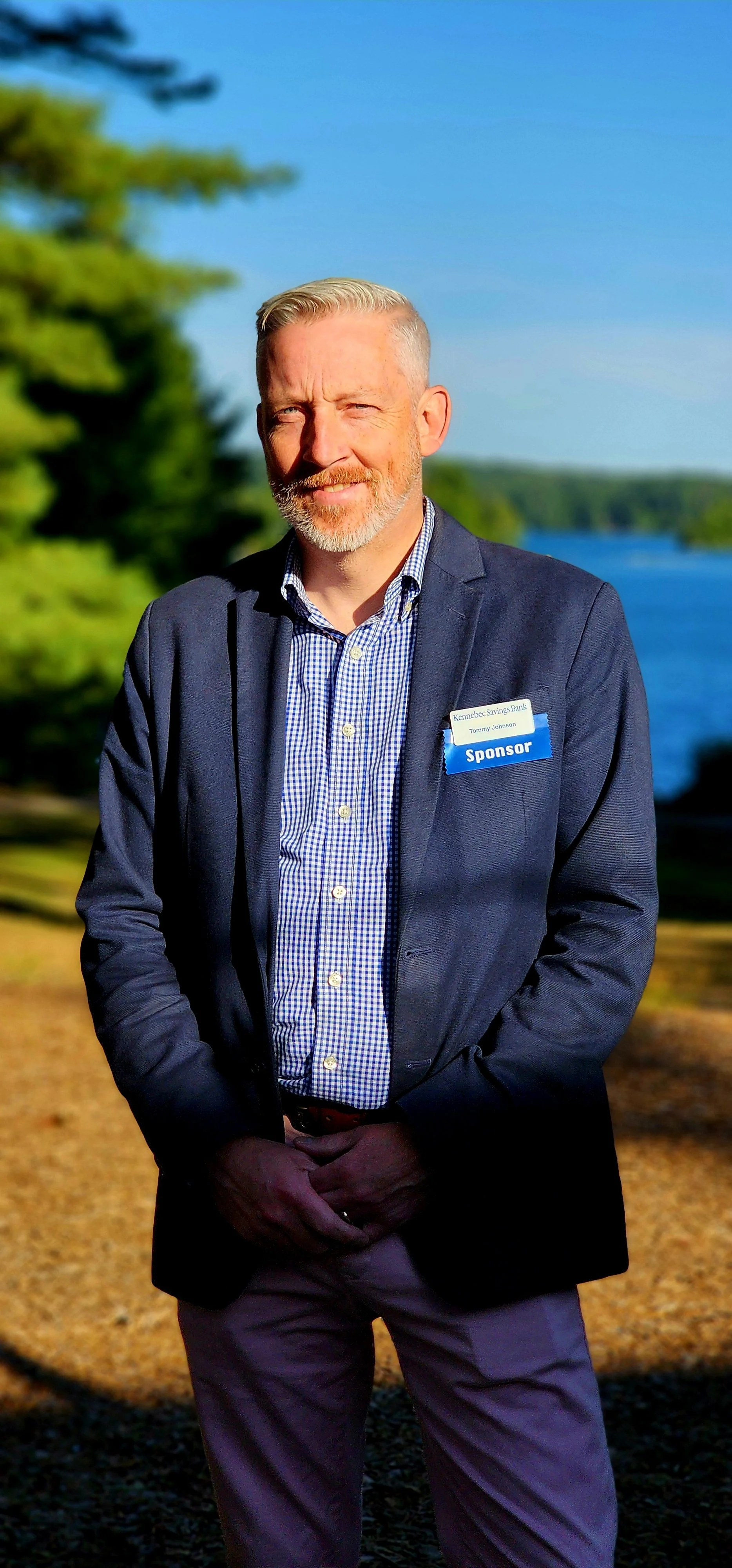 A man, wearing a navy sport coat over a blue and white checked button up shirt and khakis, facing the camera with a lake and trees in the background