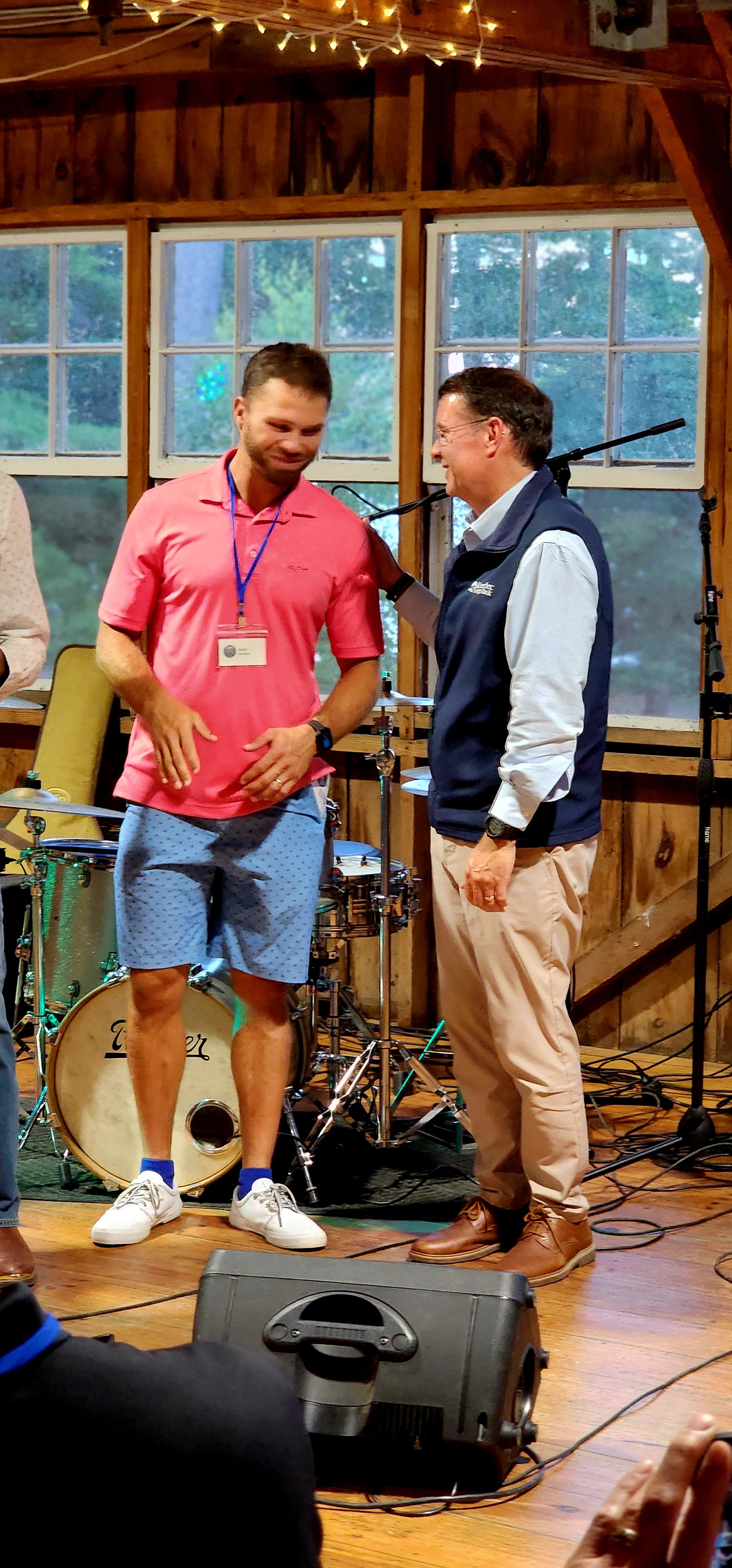 Two men, one wearing a red polo the other wearing a dark blue vest over a light blue long sleeve and khakis, standing by a microphone