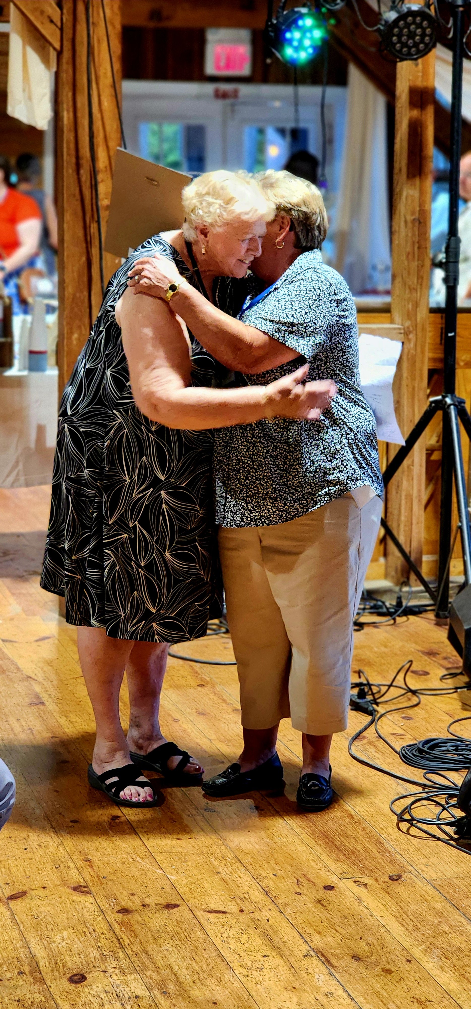 Two women, one wearing a black dress with white patterns, the other wearing a blue and white floral shirt and white pants, hugging