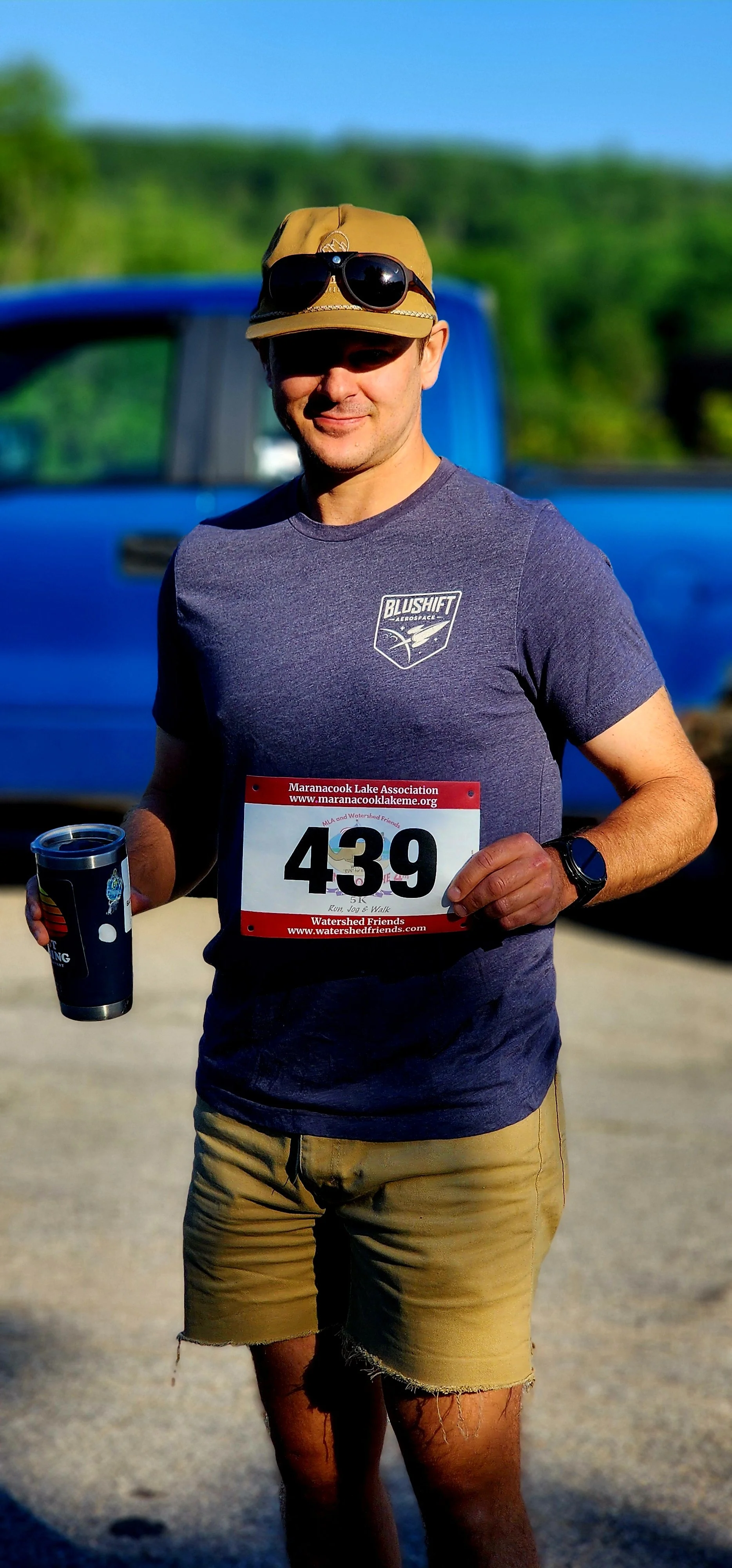 A man, wearing a grey t-shirt, tan shorts, and sunglasses over a tan ball cap,  holding a race bib number smiling at the camera