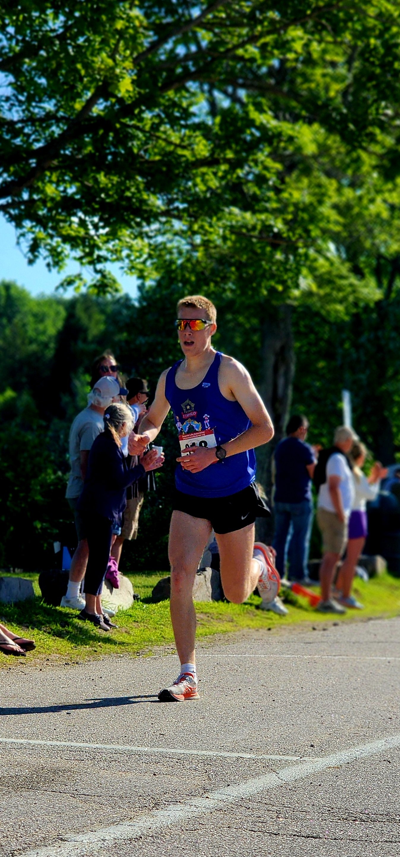 A man wearing a blue tank top and black shorts running with a crowd standing behind him