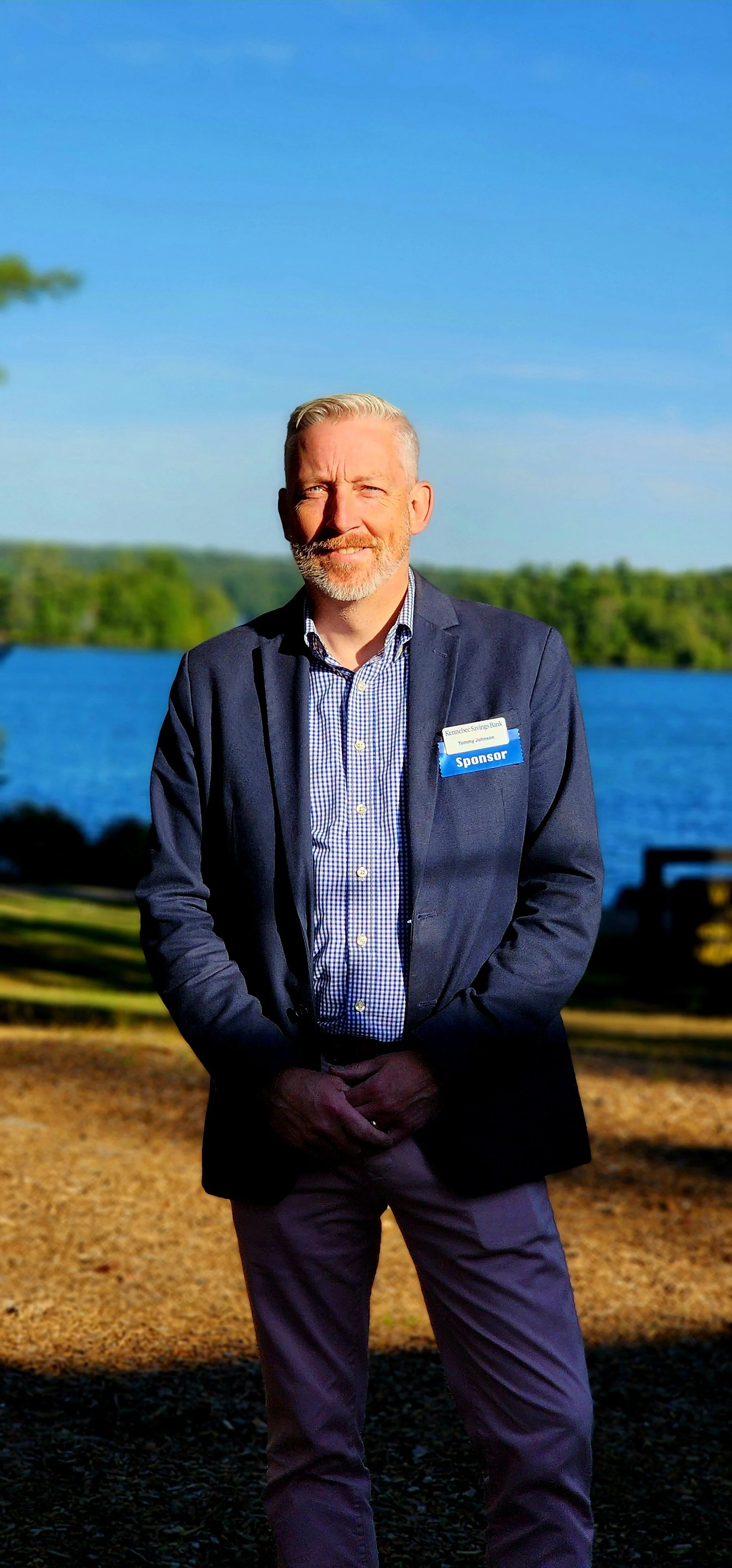 A man, wearing a navy sport coat over a blue and white checked button up shirt and khakis, facing the camera with a lake in the background
