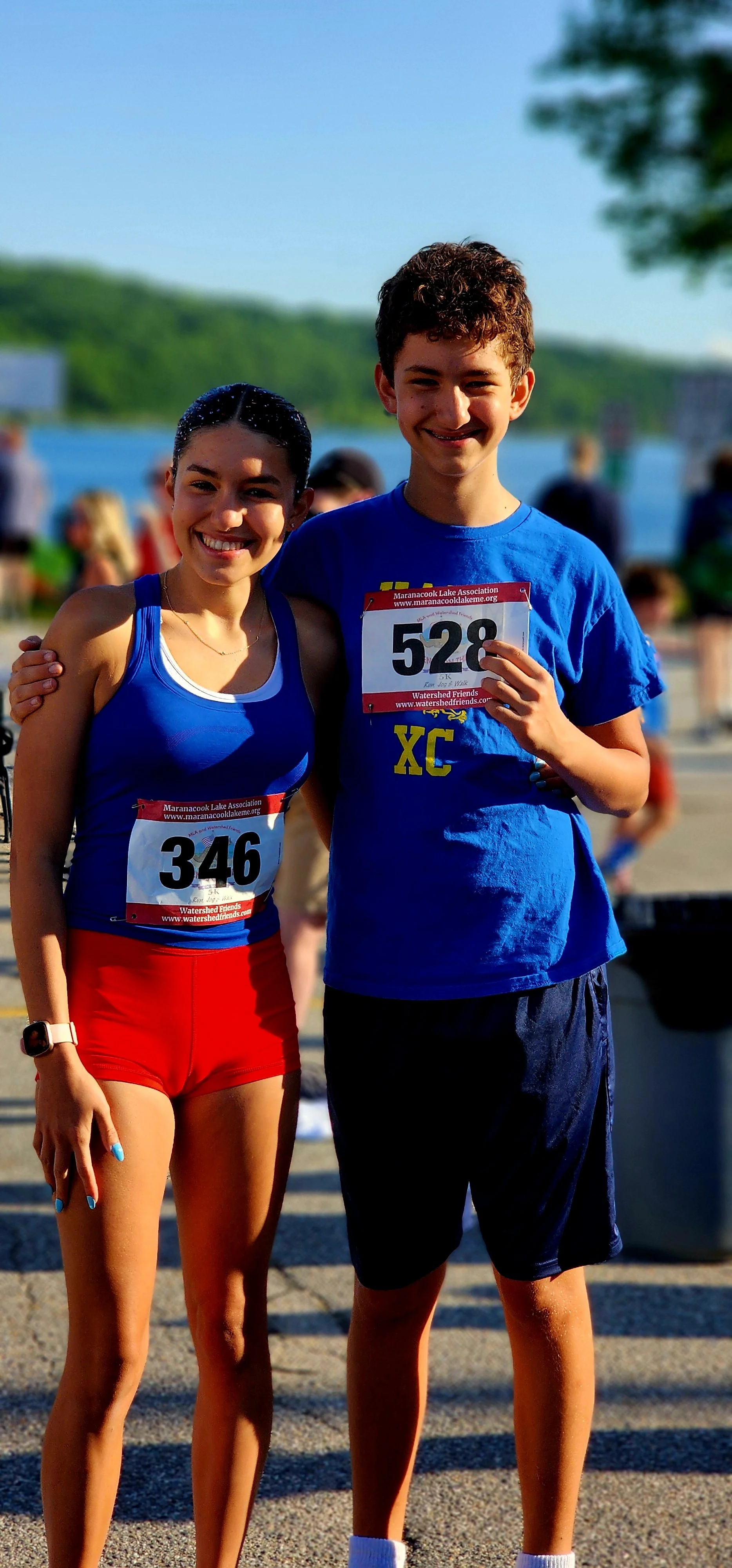 A woman, wearing a blue tank top and red shorts, and a man, wearing a blue t-shirt and black shorts, smiling at the camera, both with race bib numbers