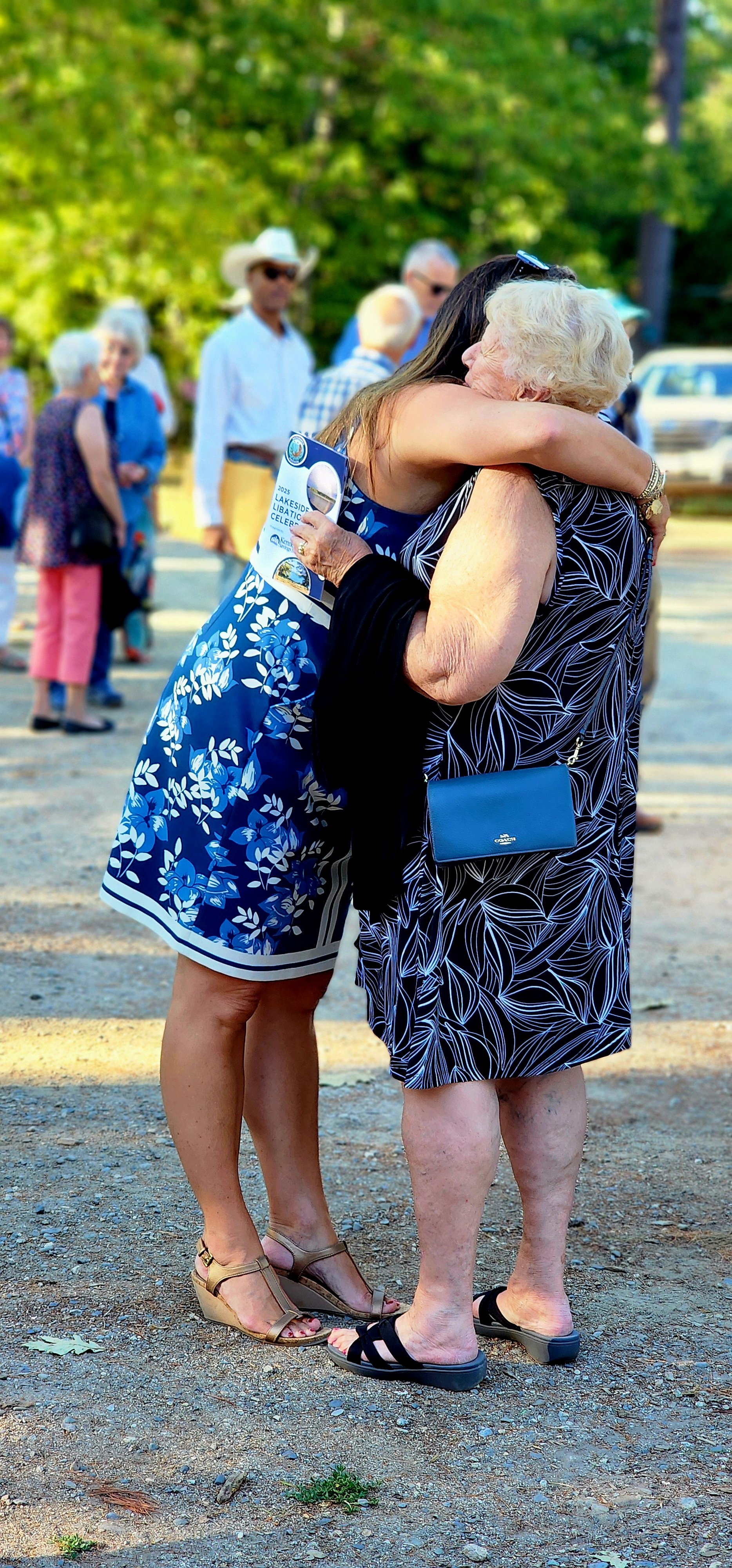 Two women, one wearing a blue floral pattern dress, the other wearing a black dress with white patterns, hugging