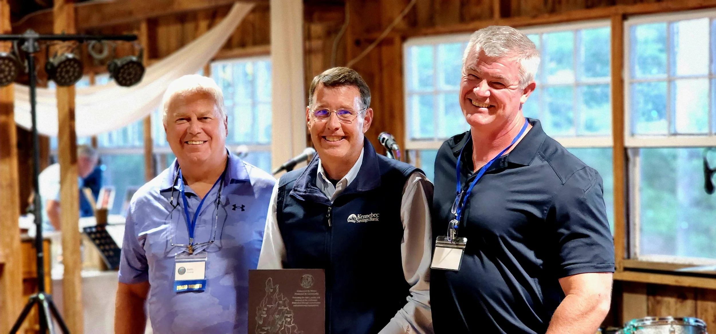 Three men smiling at the camera, the man in the middle is holding a plaque