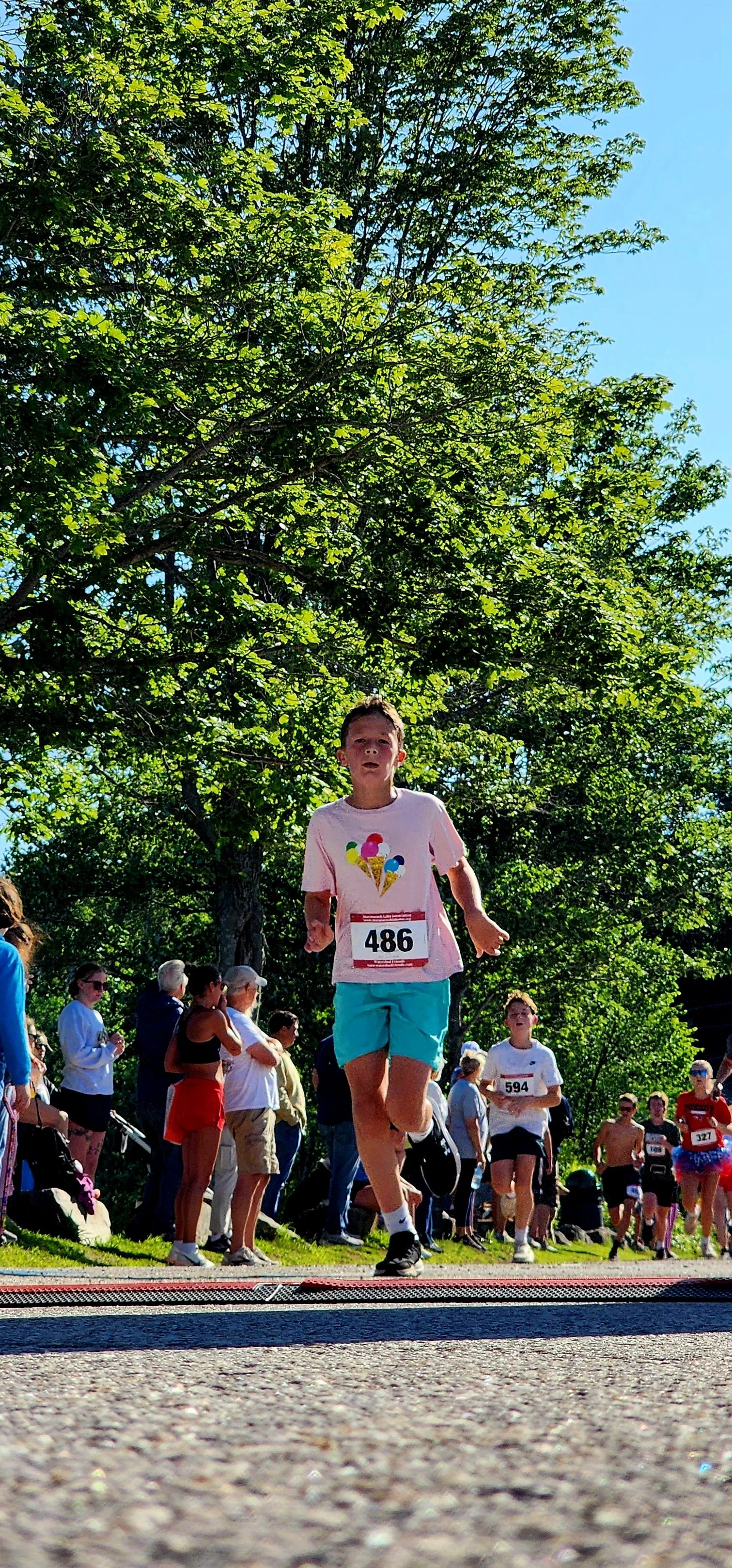 A man wearing a white t-shirt and light blue shorts running towards the finish line with a crowd running behind him
