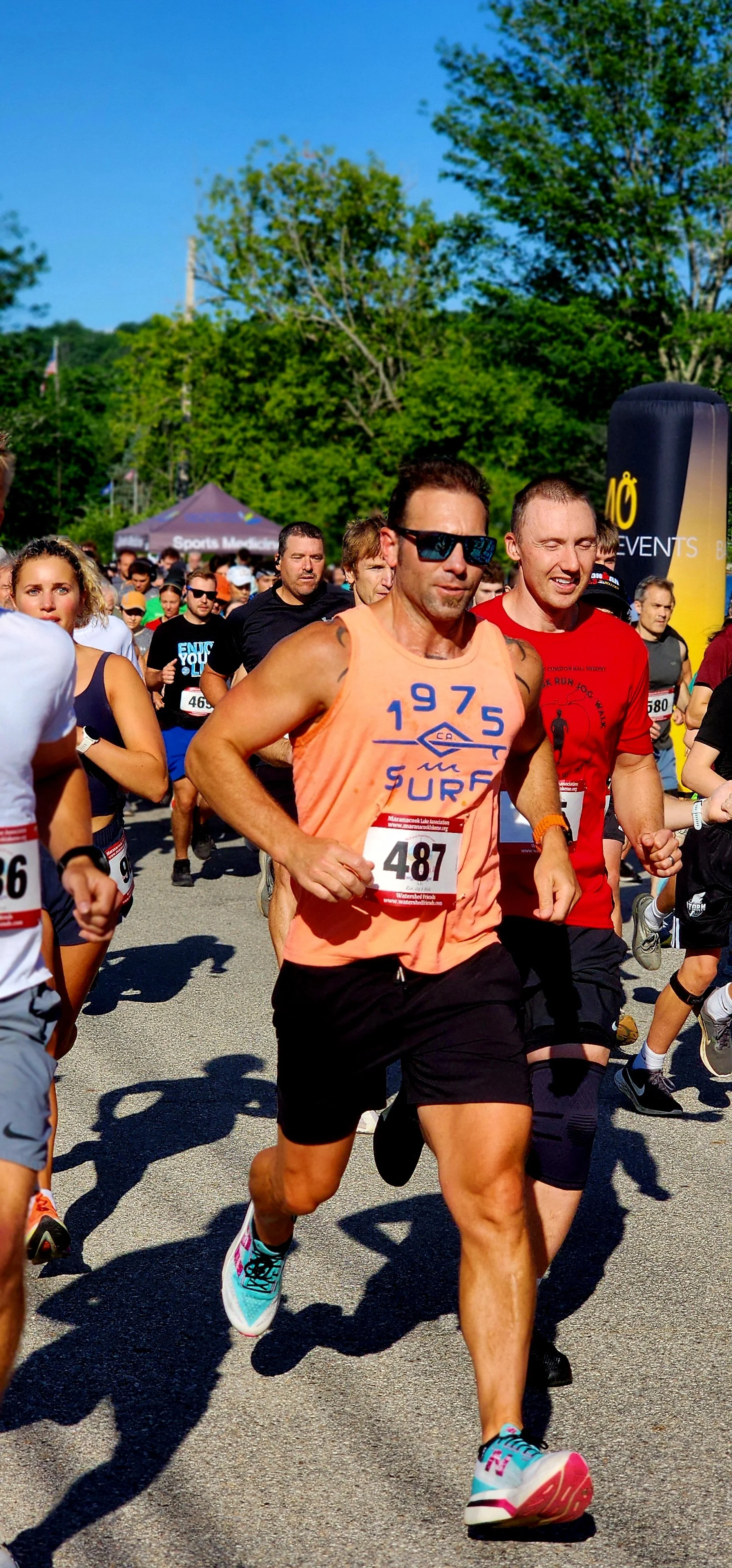 A man, wearing an orange tank top and black shorts, running with another man, wearing a red t-shirt and black shorts, running behind him