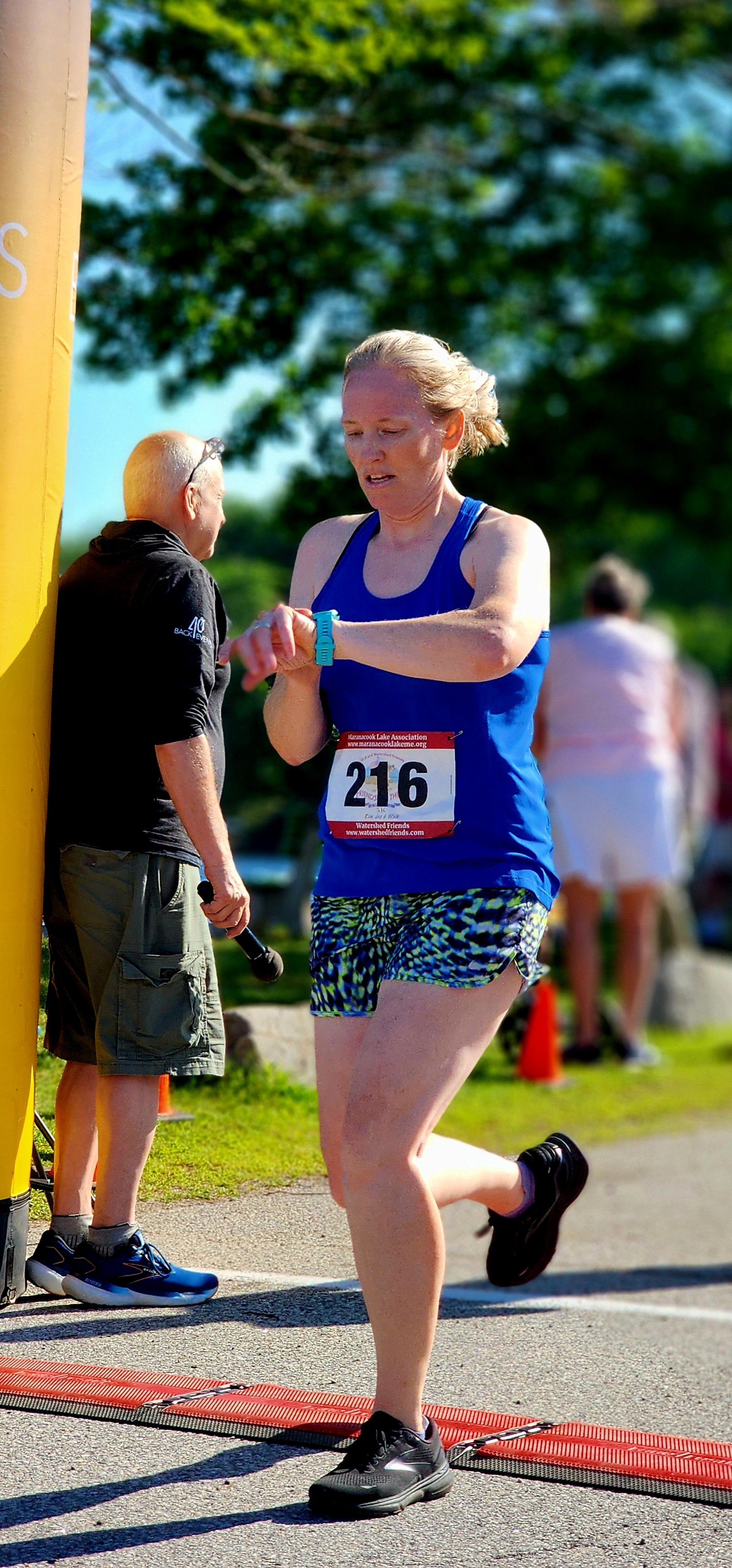 A woman wearing a blue tank top and blue green and black shorts checking her watch just past the finish line