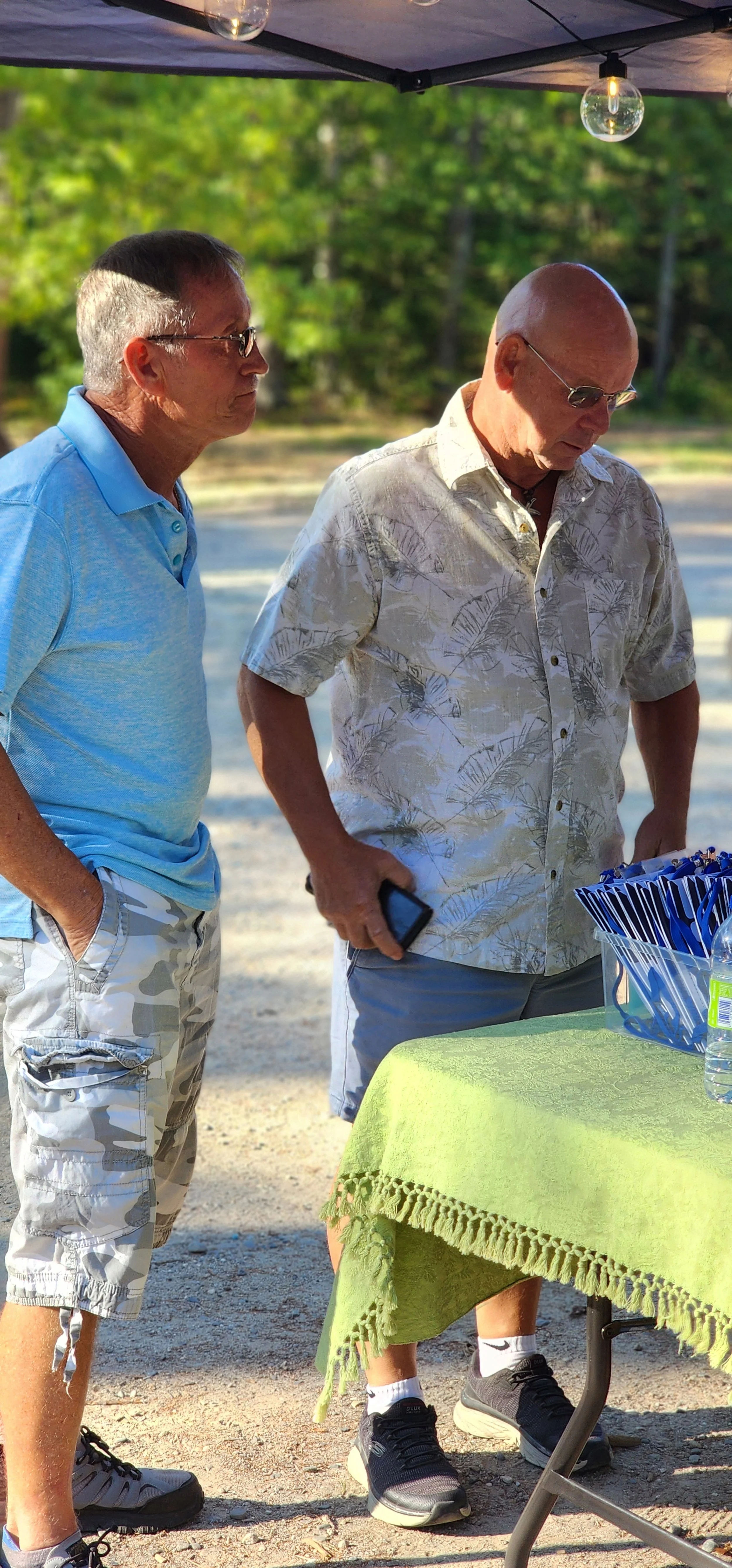 Two men, the one on the left wearing a blue polo shirt and grey camo short, the one on the right wearing a white Hawaiian shirt and grey shorts, standing at a registration camera