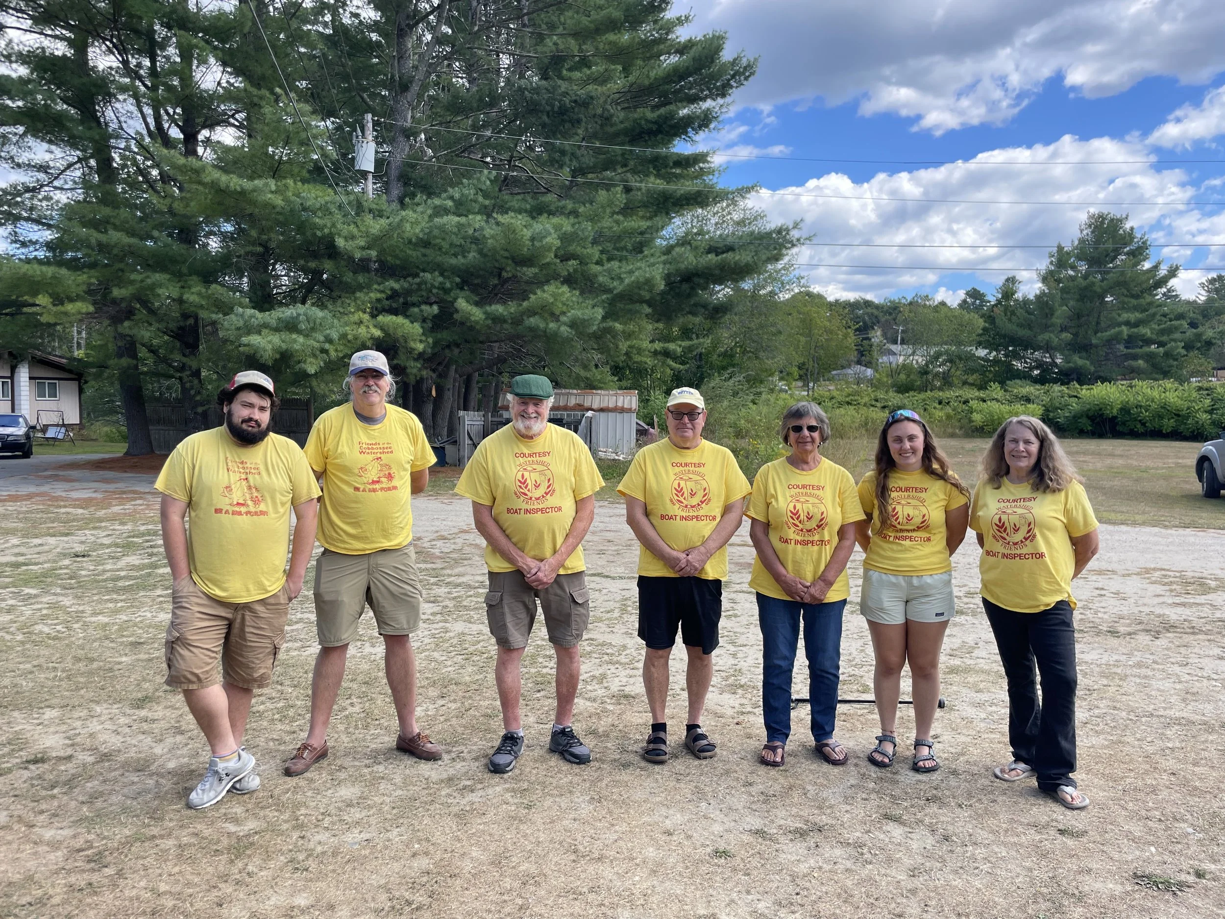 Seven people lined up smiling at the camera, all wearing yellow "Courtesy Boat Inspector" t-shirts