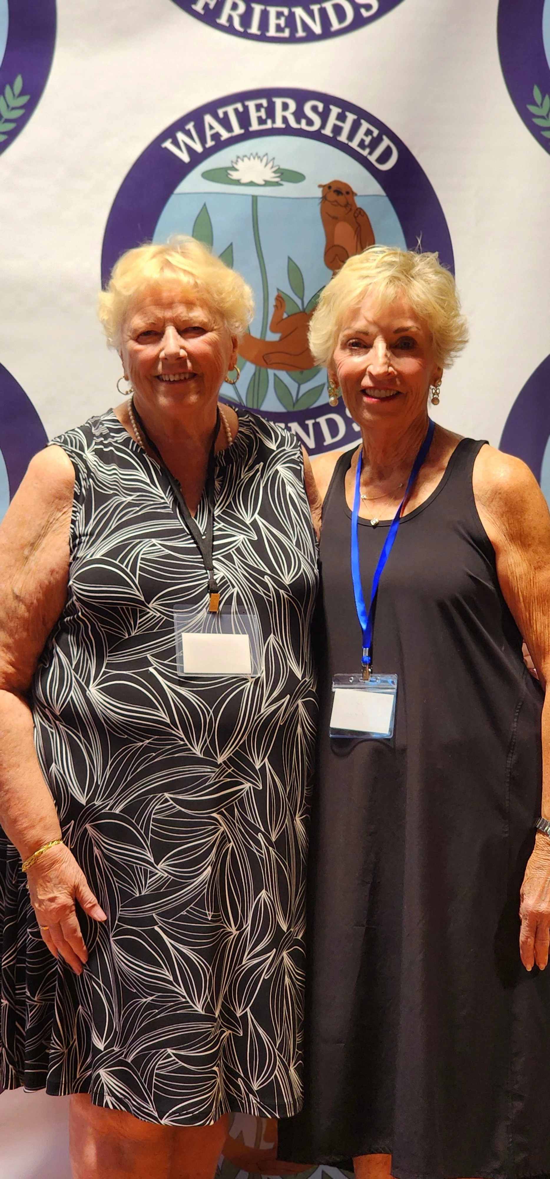 Two women, one wearing a black dress with white patterns the other wearing a black dress, smiling at the camera in front of a Watershed Friends logo backdrop