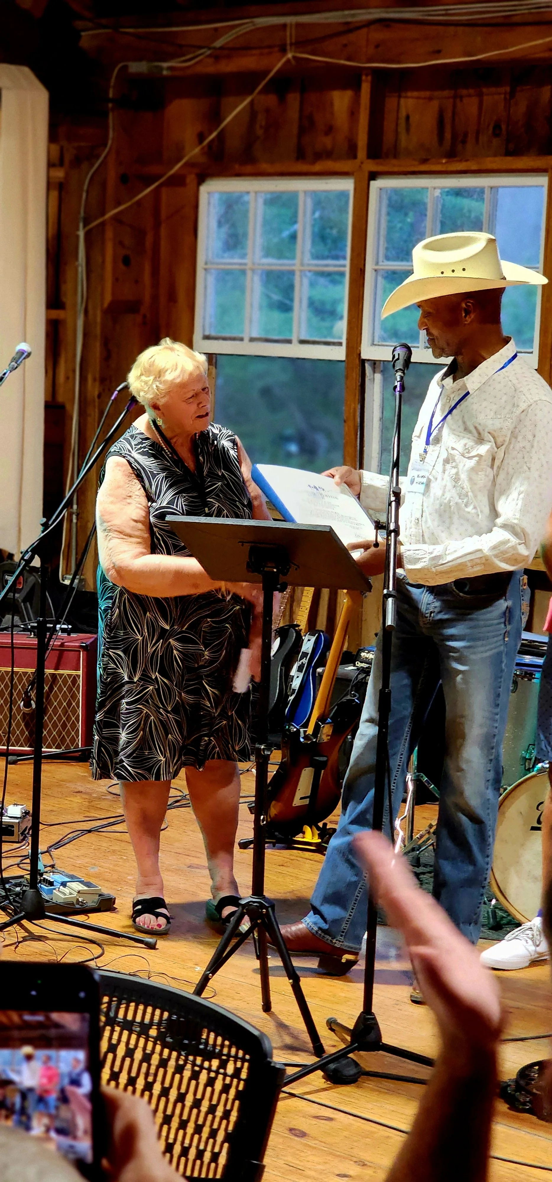 A woman, wearing a black dress with white patterns, and a man, wearing a white cowboy hat with a white shirt and jeans, the man is speaking into a microphone