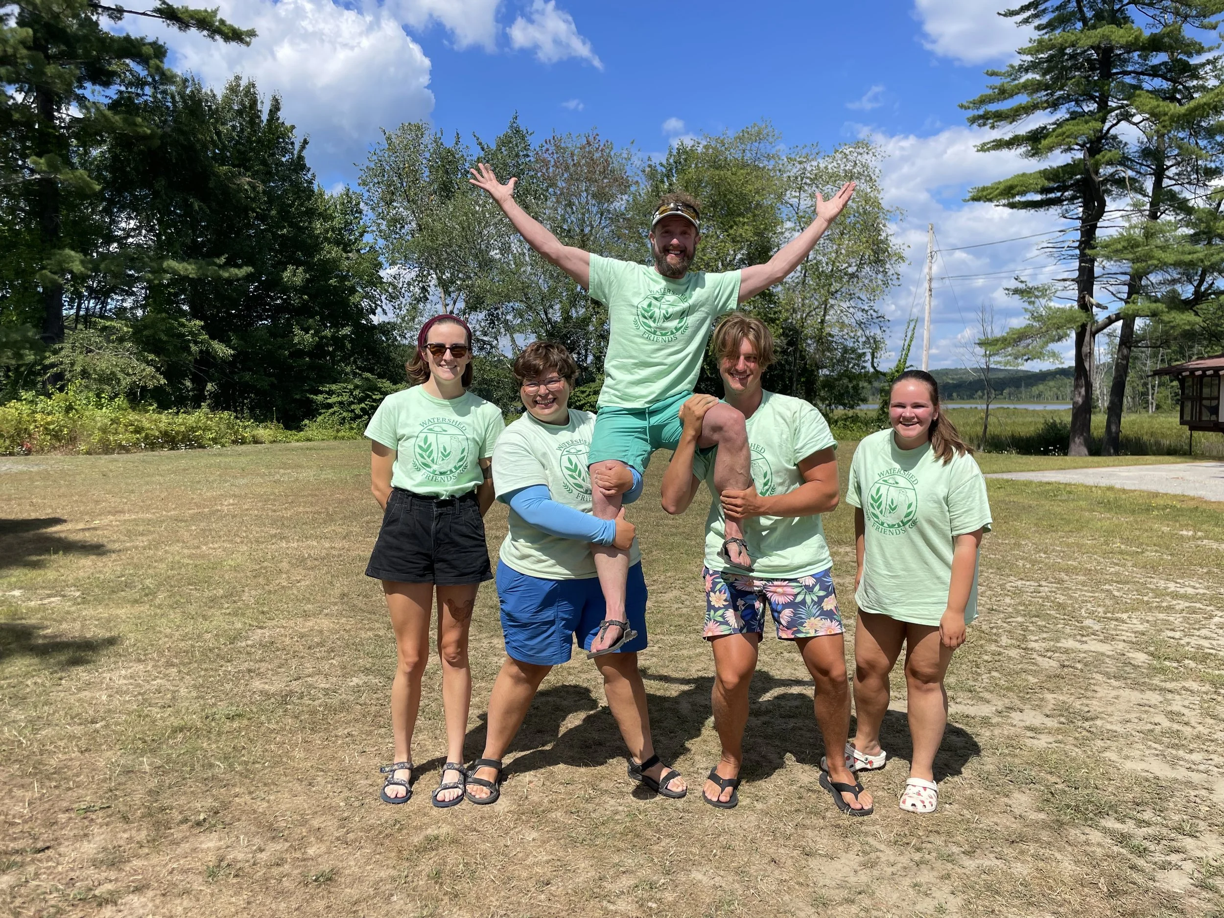 Five people facing the camera wearing matching green shirts, the one in the middle is being held in the air by the two people to either side