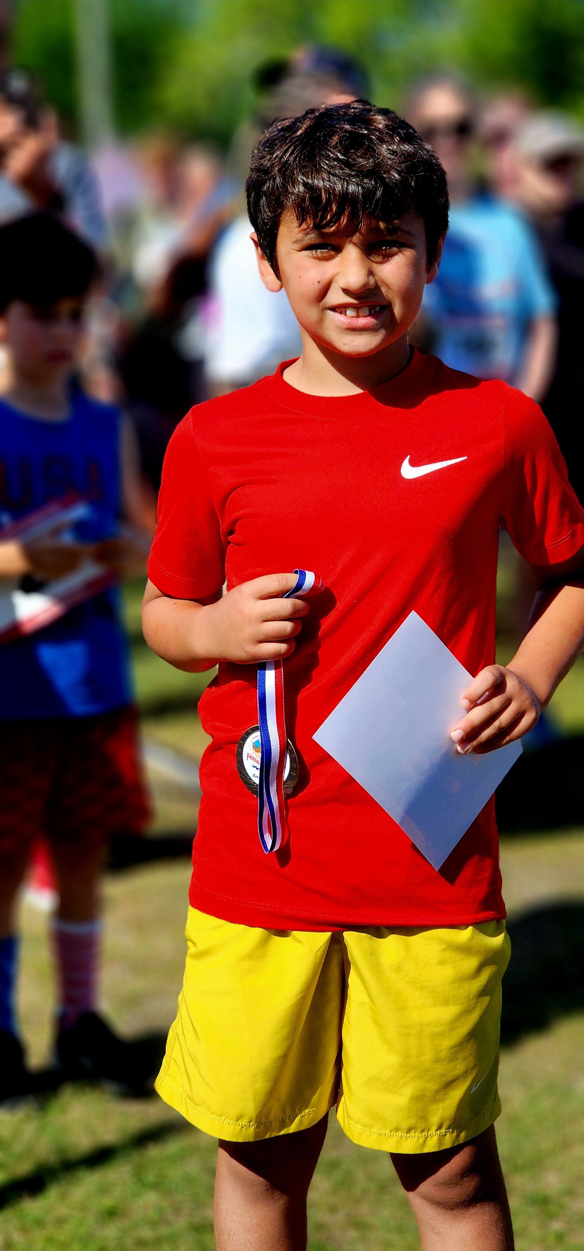 A boy wearing a red t-shirt and yellow shorts, holding a prize medal and piece of paper