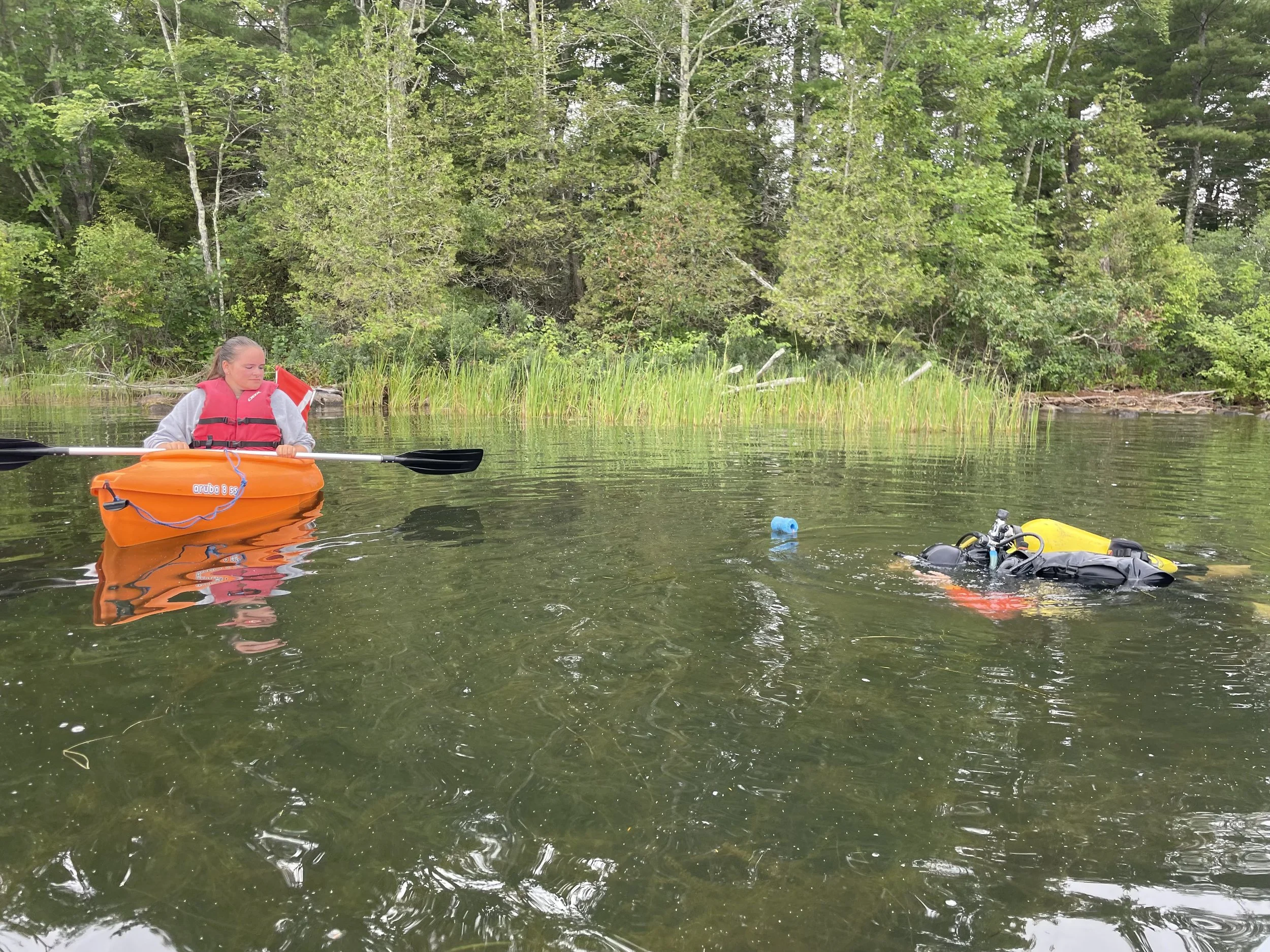 A person in an orange kayak watching a diver, who is visible just under the surface of the water