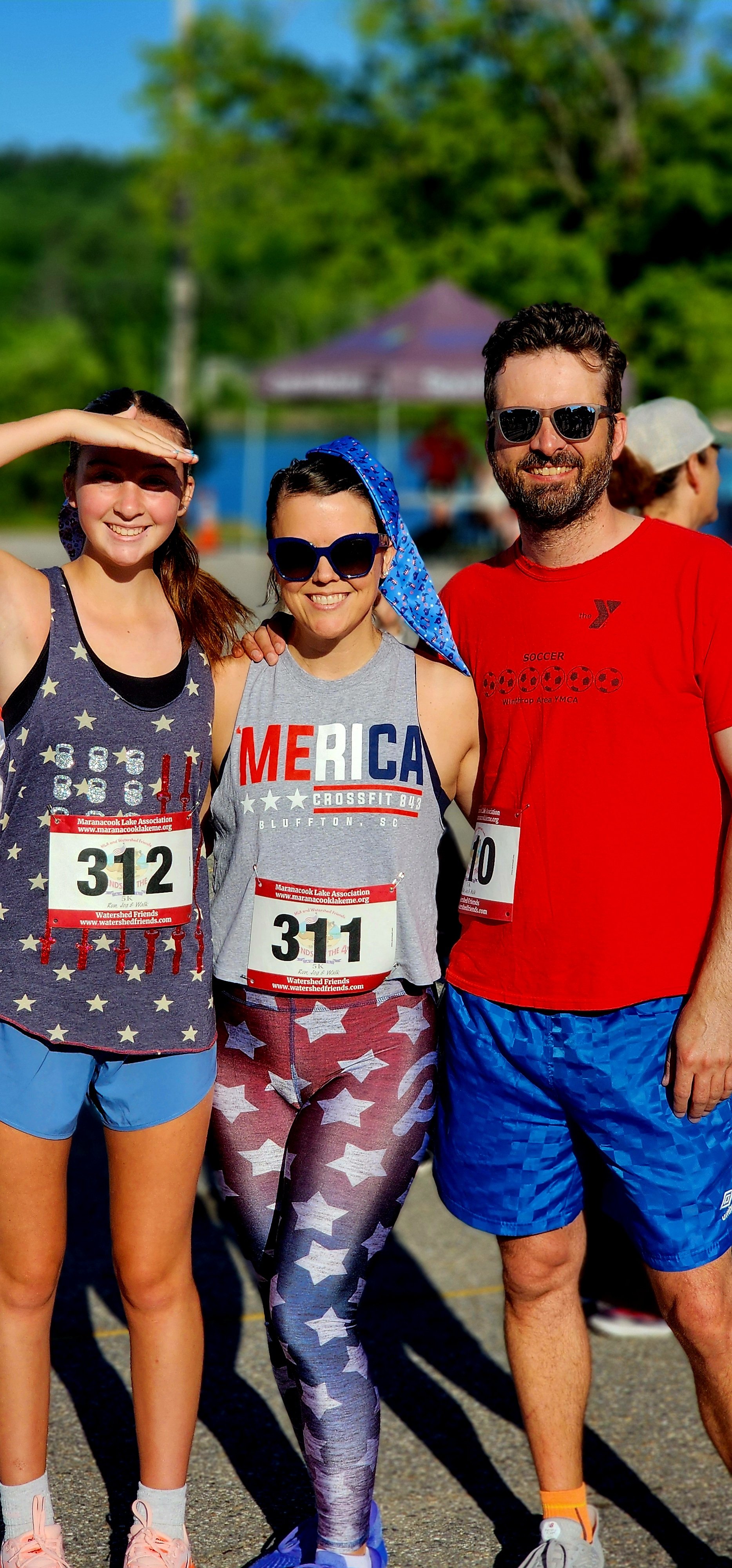 Three people, one wearing a blue tank top with the American flag and blue shirts, one wearing a grey tank top that says "America" and star printed blue and red leggings, and the third wearing a red t-shirt and blue shorts, smiling at the camera weari