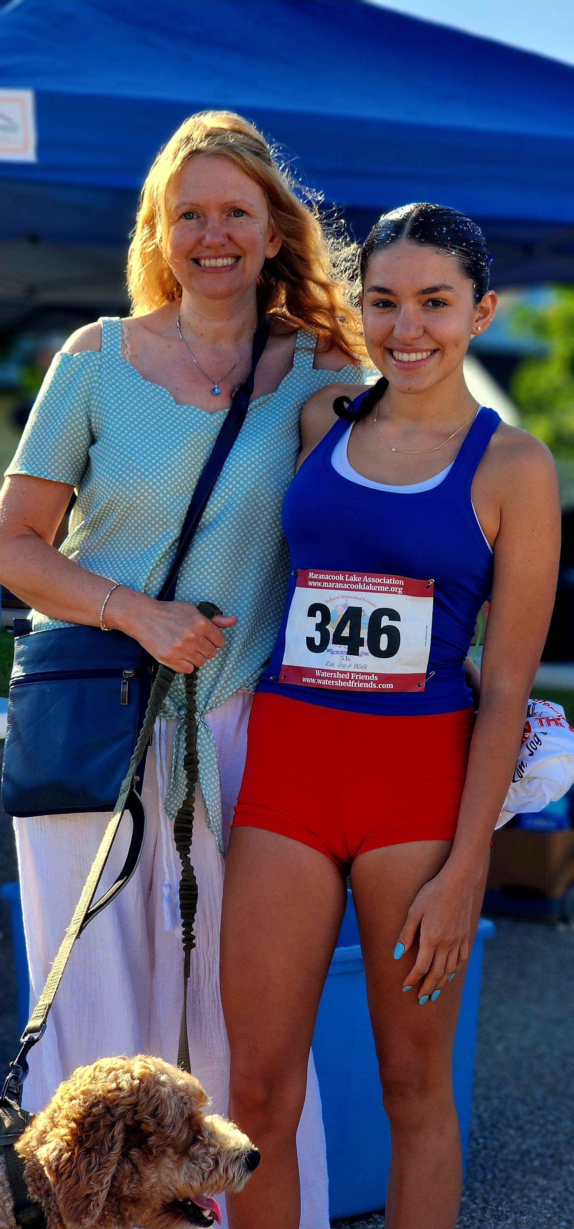 Two women smiling at the camera, the one on the left holding the leash of a dog and wearing a blue t-shirt with white skirt and blue crossbody bag and the one on the right wearing a bib number with red shorts and a blue tank top 