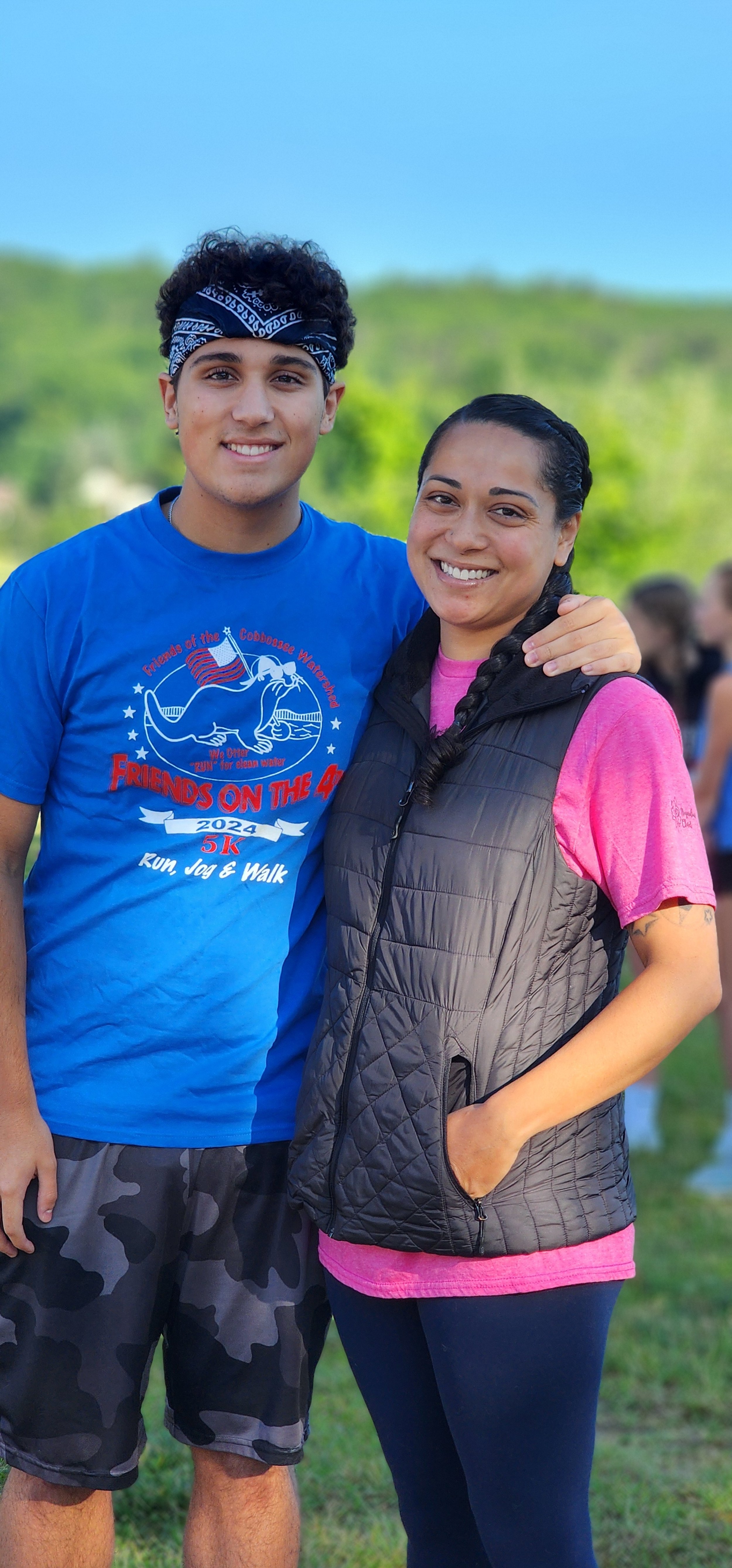 A man, wearing a blue t-shirt and grey camo patterned shorts, and a woman, wearing a dark grey vest over a pink t-shirt and navy leggings, smiling at the camera