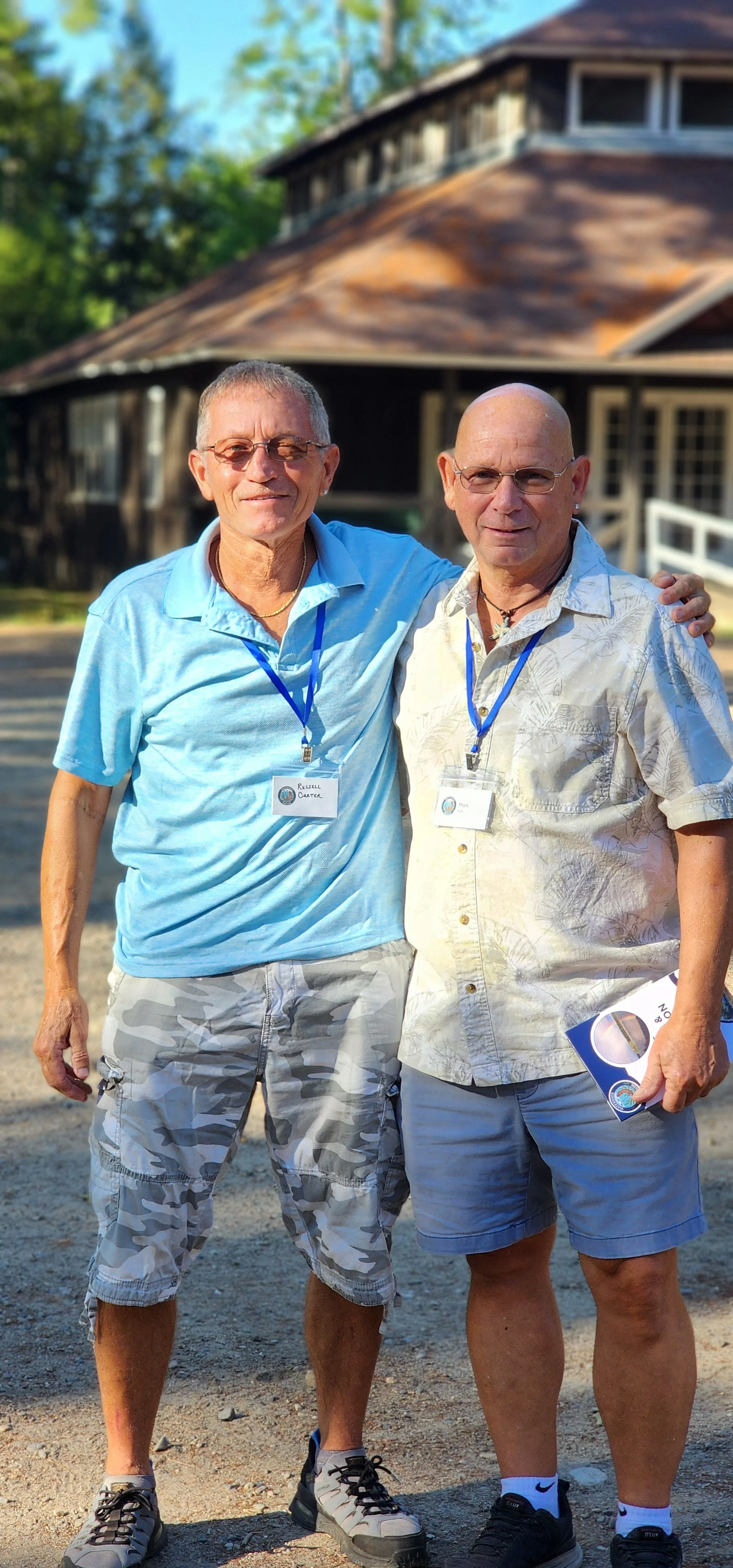 Two men, to one on the left wearing a blue polo shirt and grey camo and the one on the right wearing a white Hawaiian shirt and grey shorts, smiling at the camera