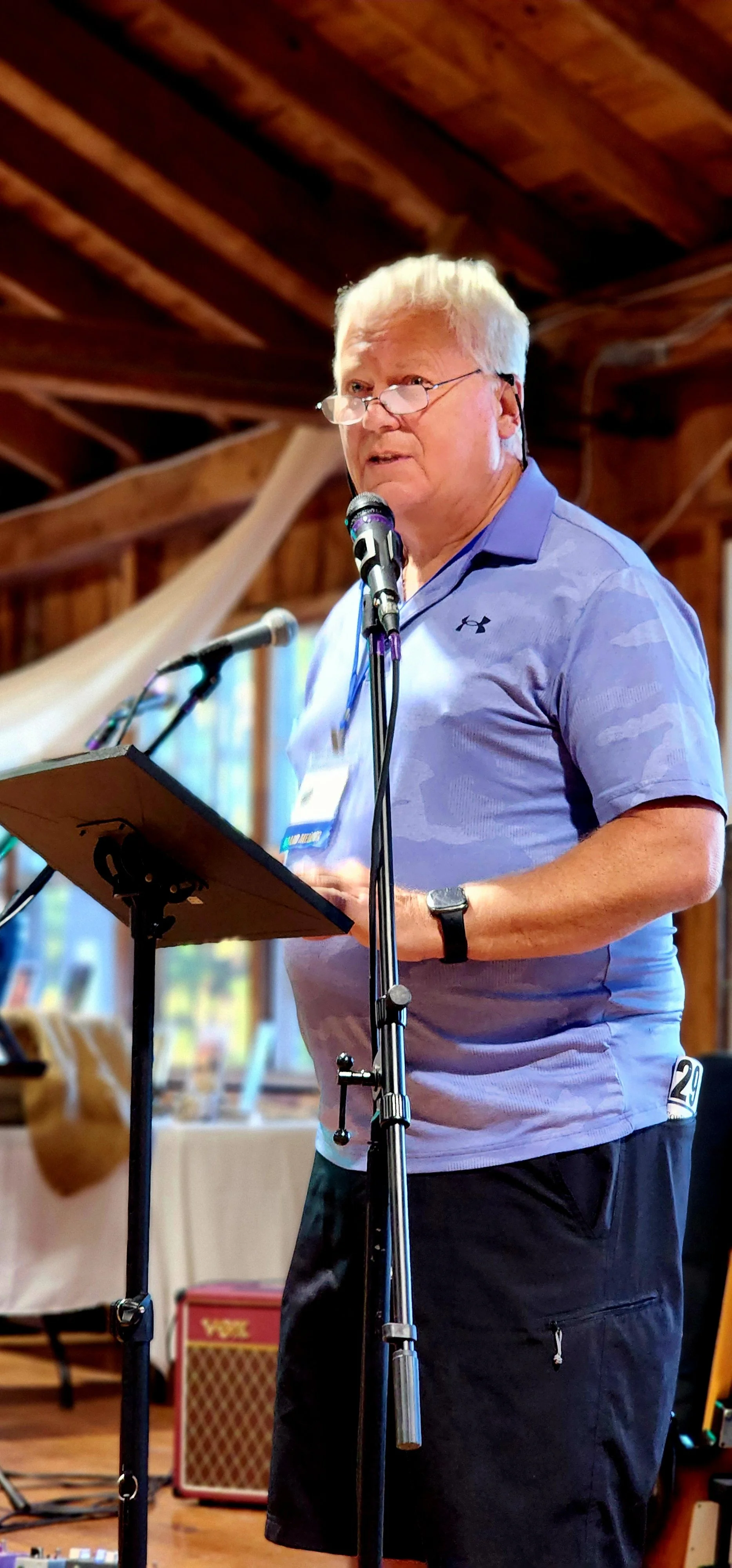A man, wearing a light blue polo and black shorts, speaking into a microphone with a stand in front of him
