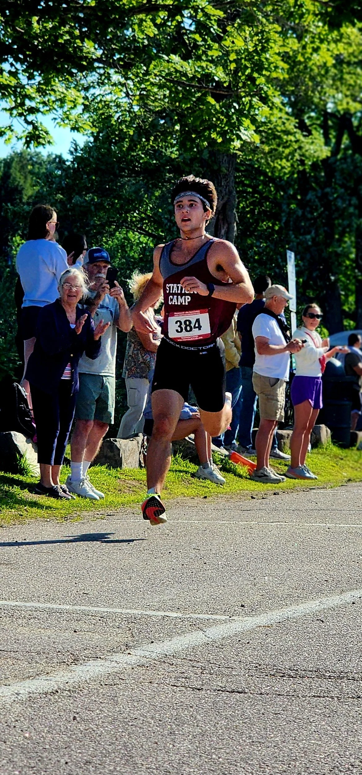 A man wearing a grey and maroon tank top and black shorts running with a crowd standing behind him