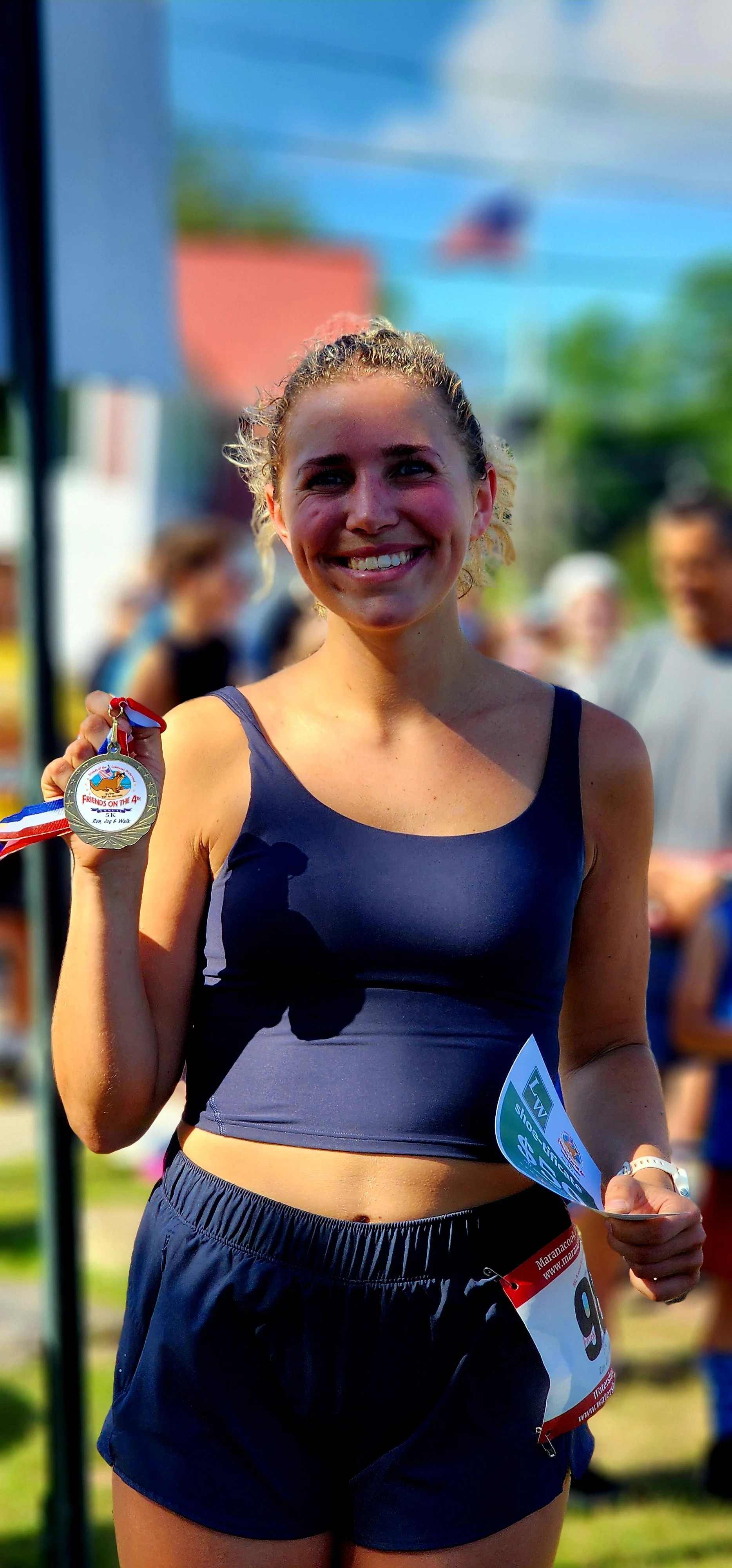 A woman smiling at the camera, wearing a blue tank top and shorts holding up an award medal