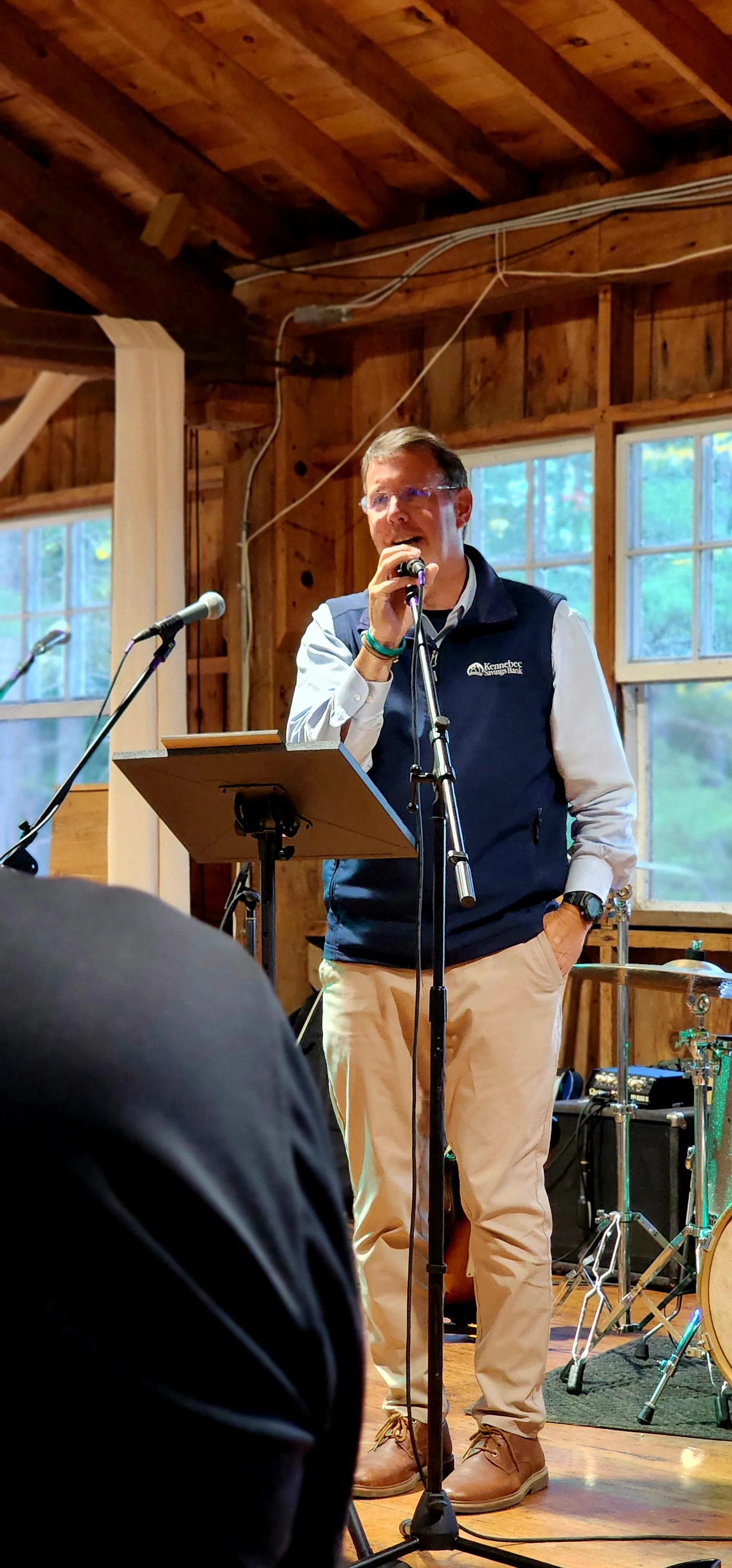 A man, wearing a dark blue vest over a light blue long sleeve and khakis, speaking into a microphone with a stand in front of him