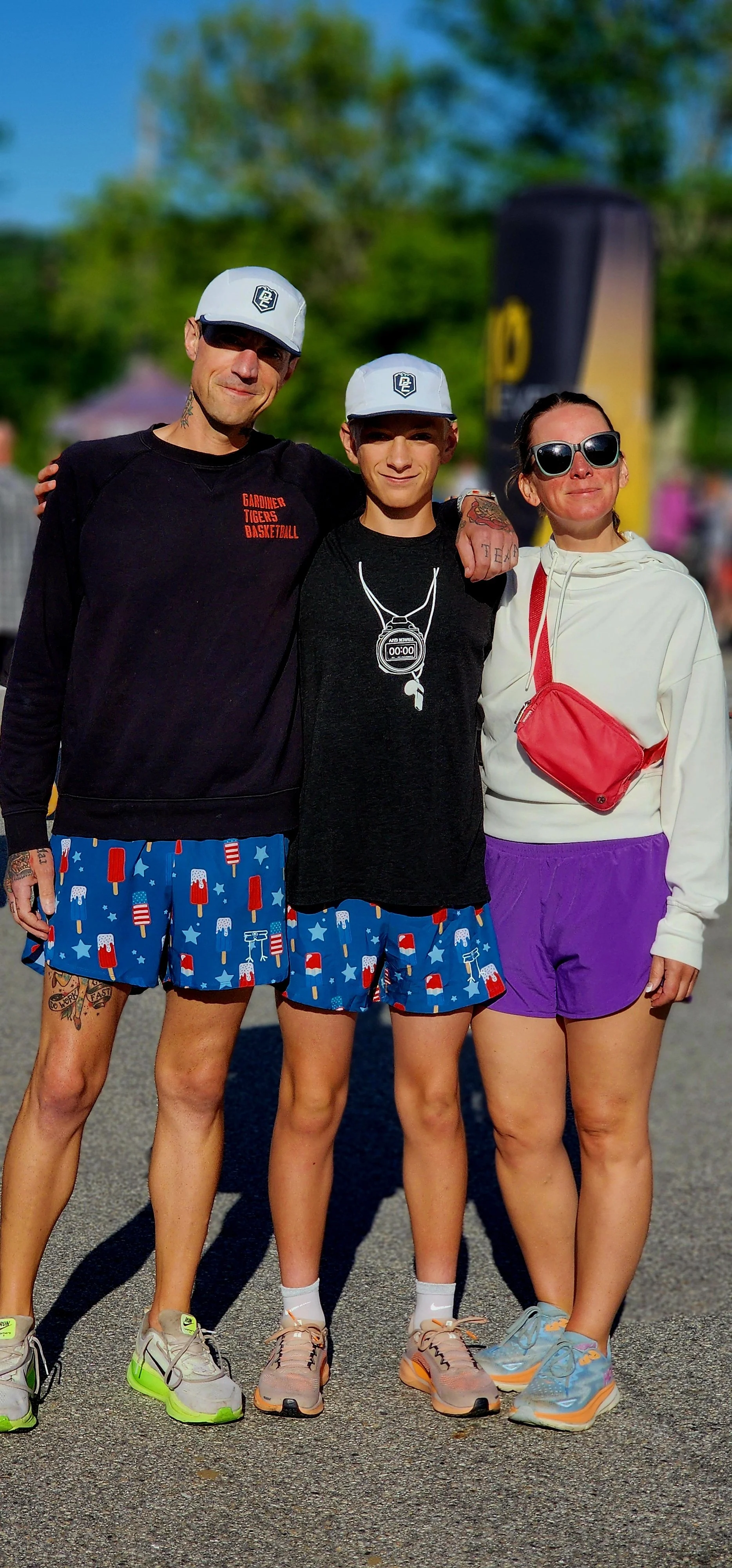Three people, two men wearing black long sleeve shirts and matching blue shorts and a woman wearing a white long sleeve shirt with purple shorts and a red cross body bag, smiling at the camera