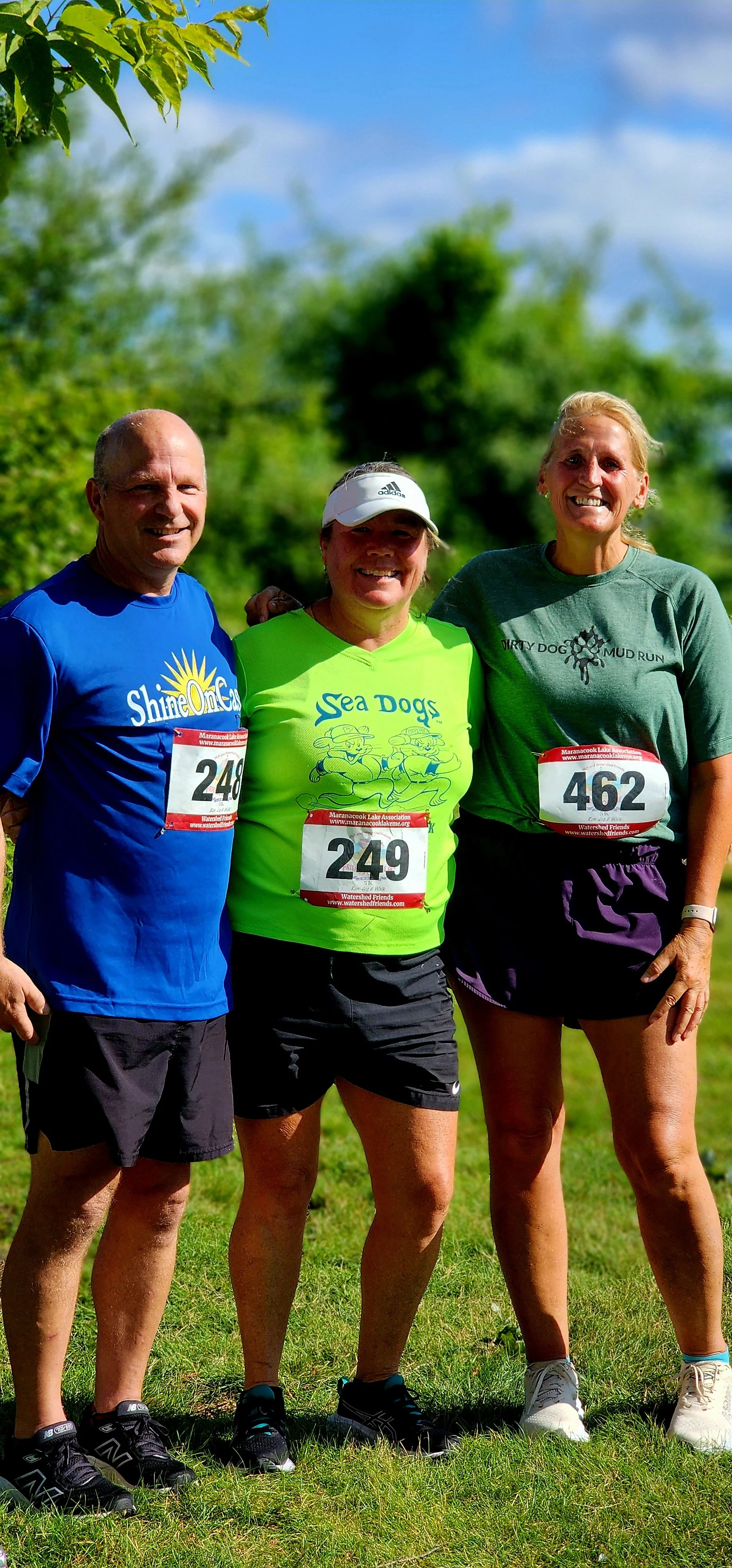 Three people smiling at the camera, the man on the left wearing a blue t-shirt and black shorts, the woman in the middle wearing a neon green shirt and black shorts, and the woman on the right wearing a green t-shirt and black shorts