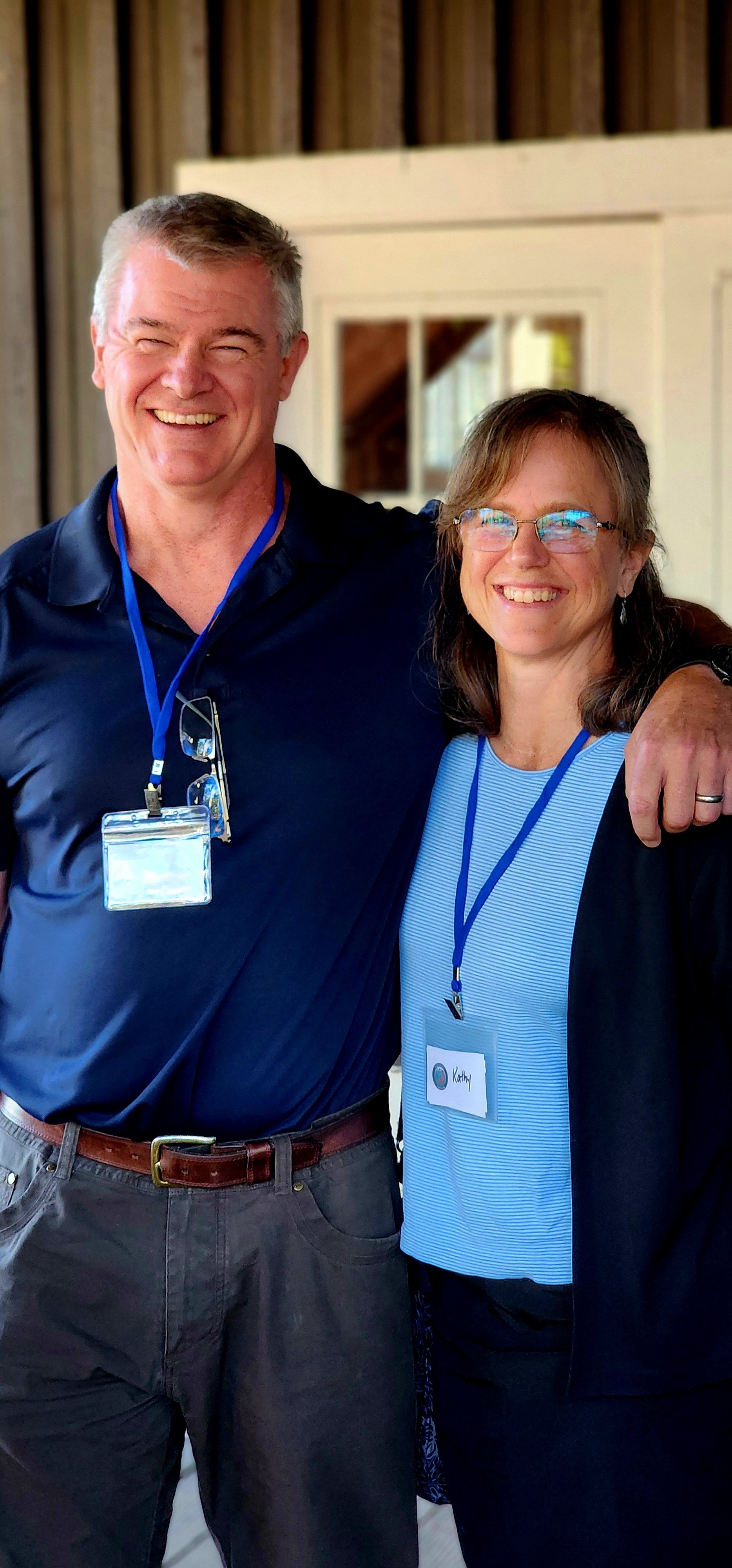 A man, wearing a navy polo and grey pants, and a woman, wearing a black cardigan over a blue shirt and black pants, smiling at the camera