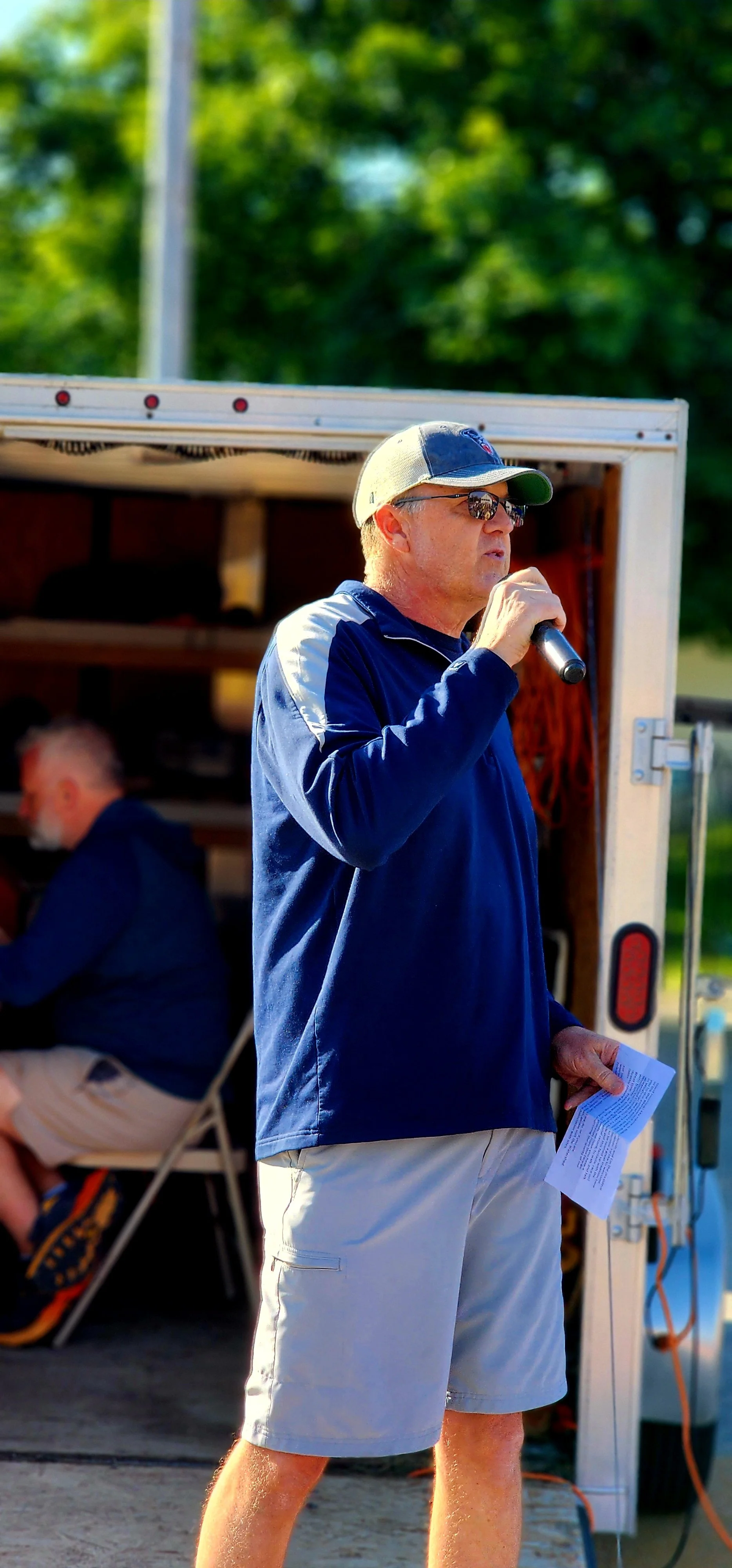 A man, wearing a blue ball cap, blue long-sleeved shirt and grey shorts, speaking into a microphone