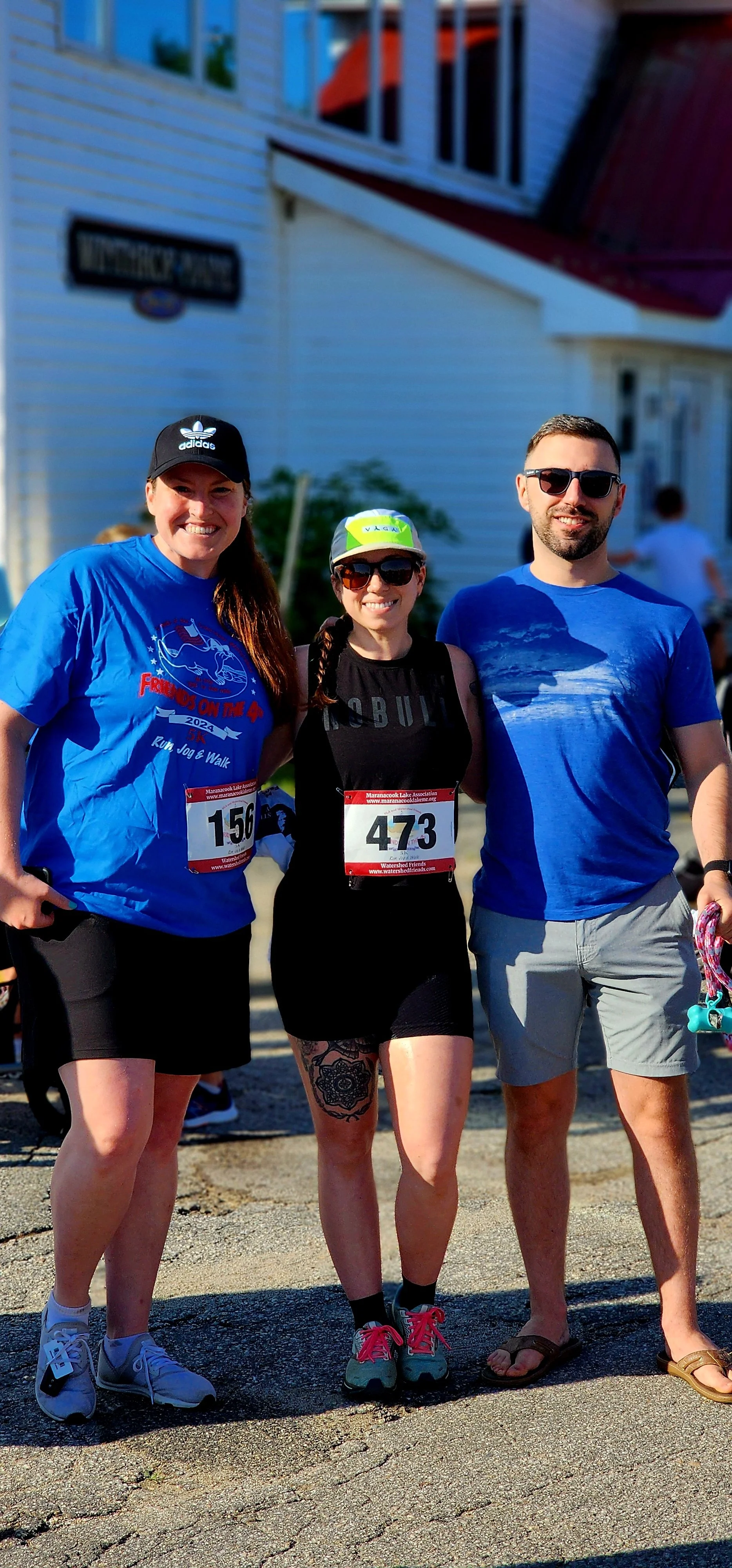Three people, one woman wearing a dark blue ball cap, blue t-shirt, and black shorts, one woman wearing a black tank top and black shorts, and a man wearing a blue t-shirt and grey shorts smiling at the camera