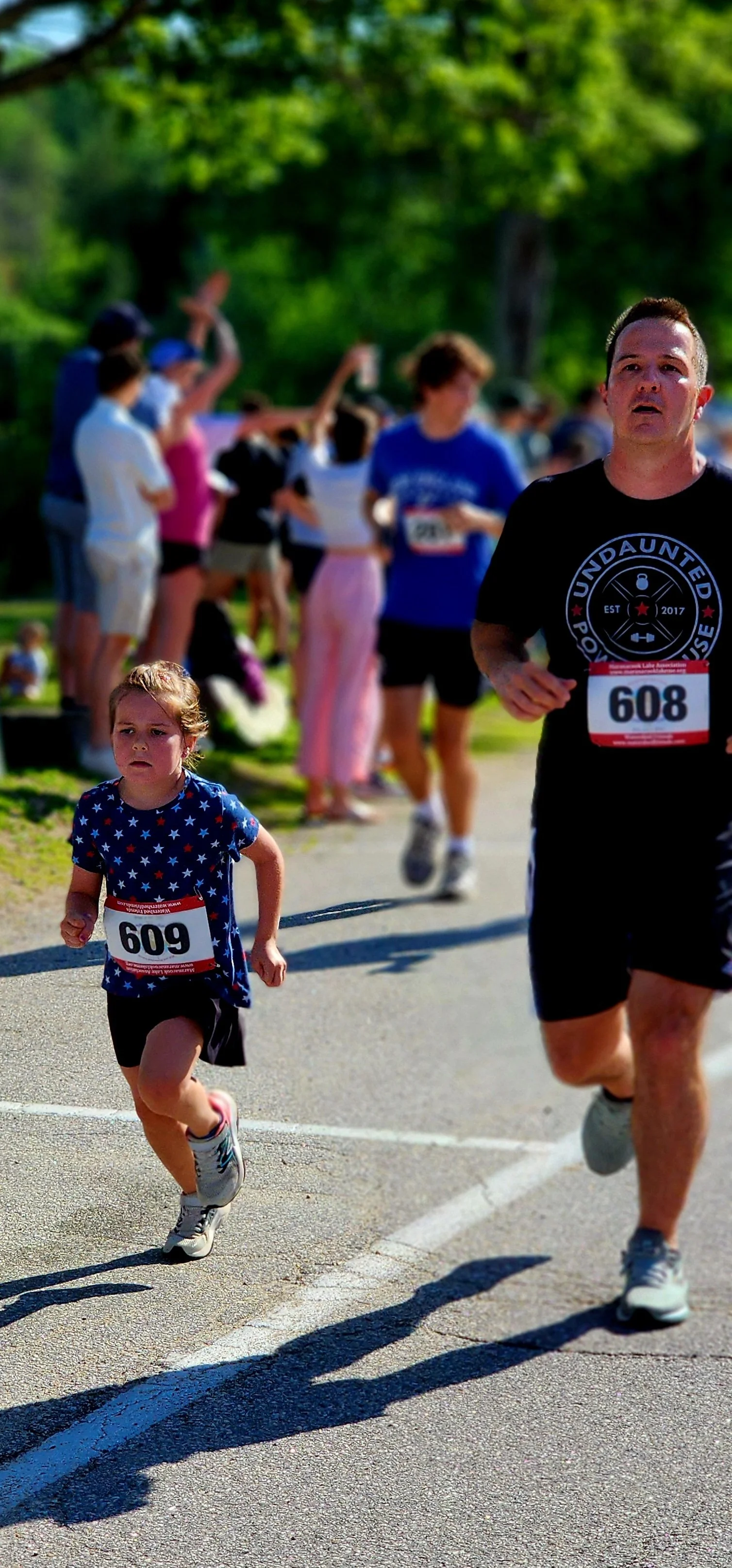 A young girl wearing a blue t-shirt with white polka dots and black shorts running alongside a man wearing a black t-shirt and black shorts