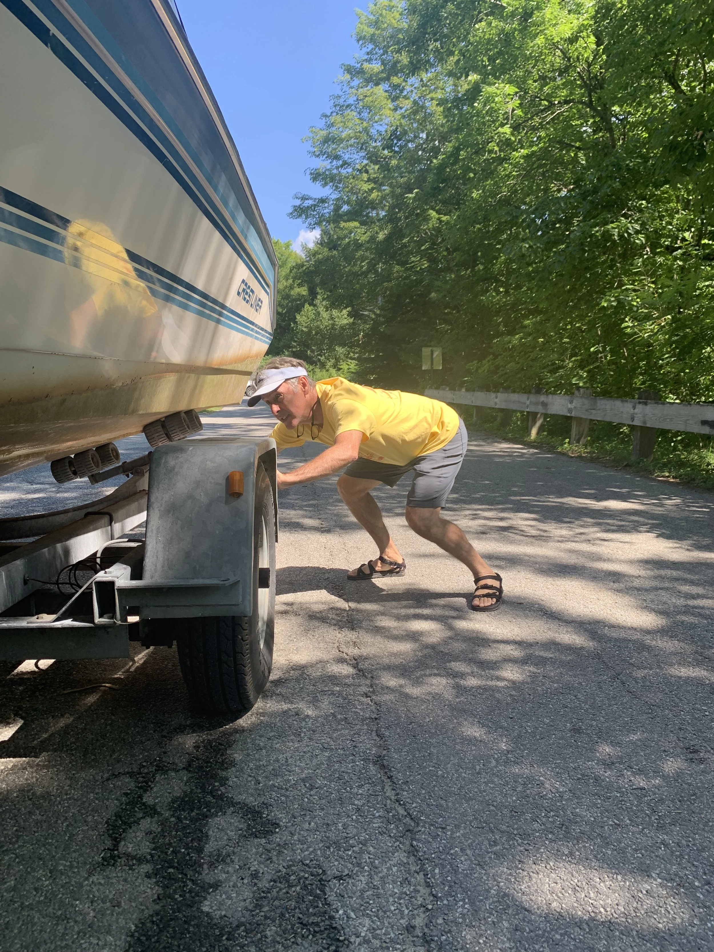 A man, wearing a yellow t-shirt, leaning forward to peer under a boat that is on a trailer