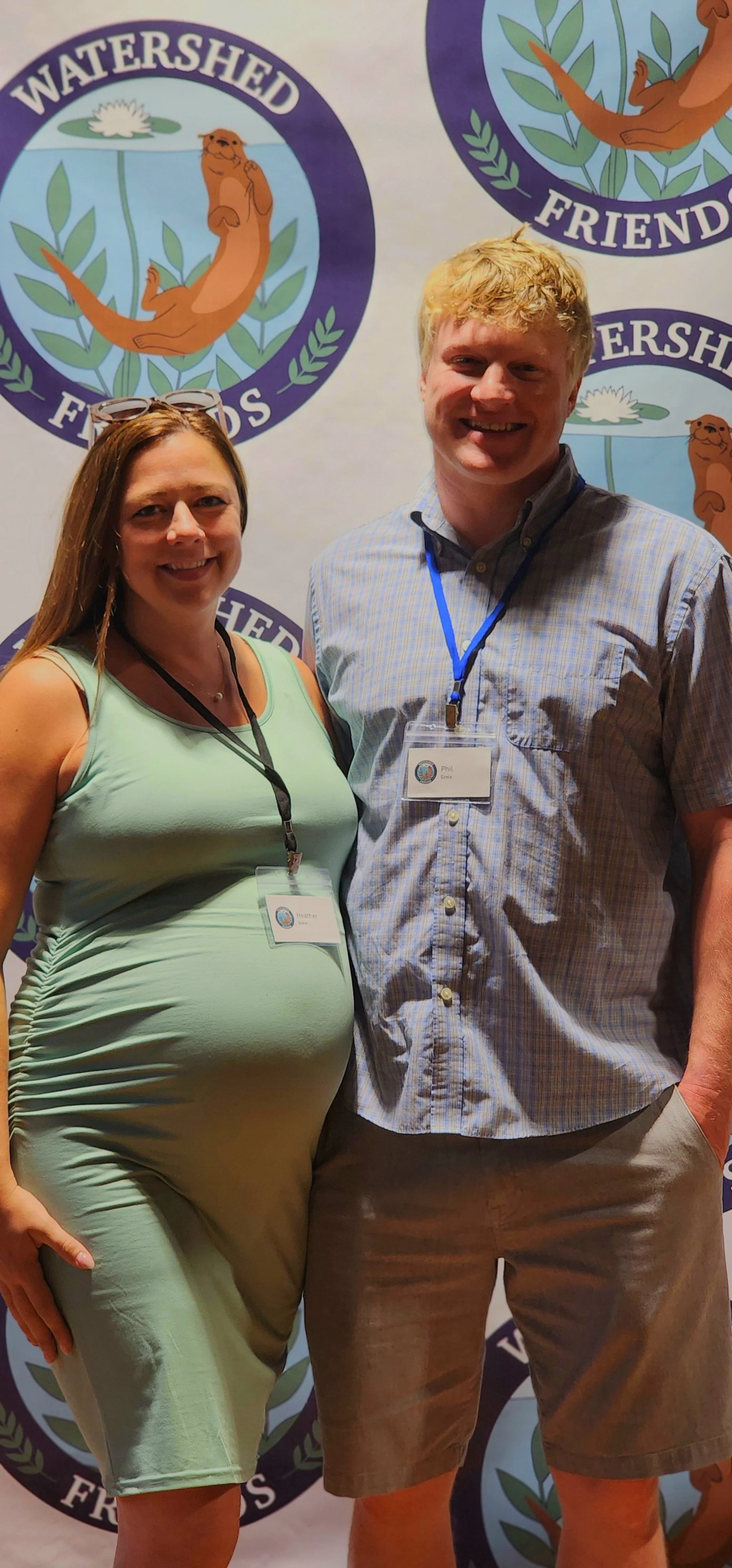 A woman, wearing a mint green sleeveless dress, and a man, wearing a short sleeve blue button up and khaki shorts, smiling at the camera