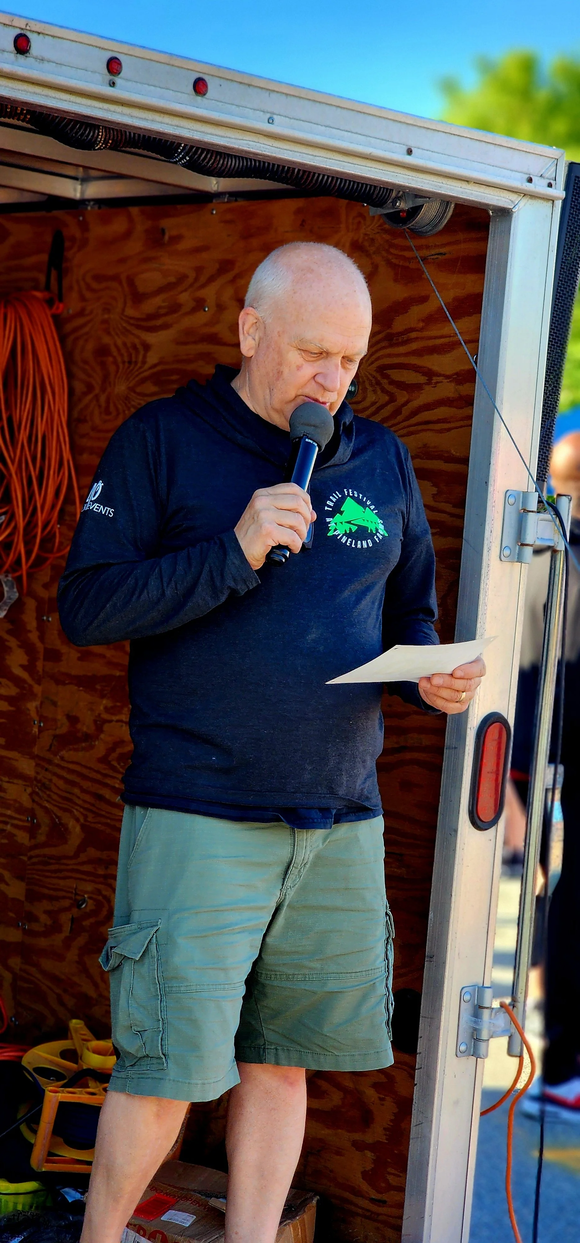 A man, wearing a black long sleeve shirt and green shorts, speaking into a microphone and reading off a sheet of paper