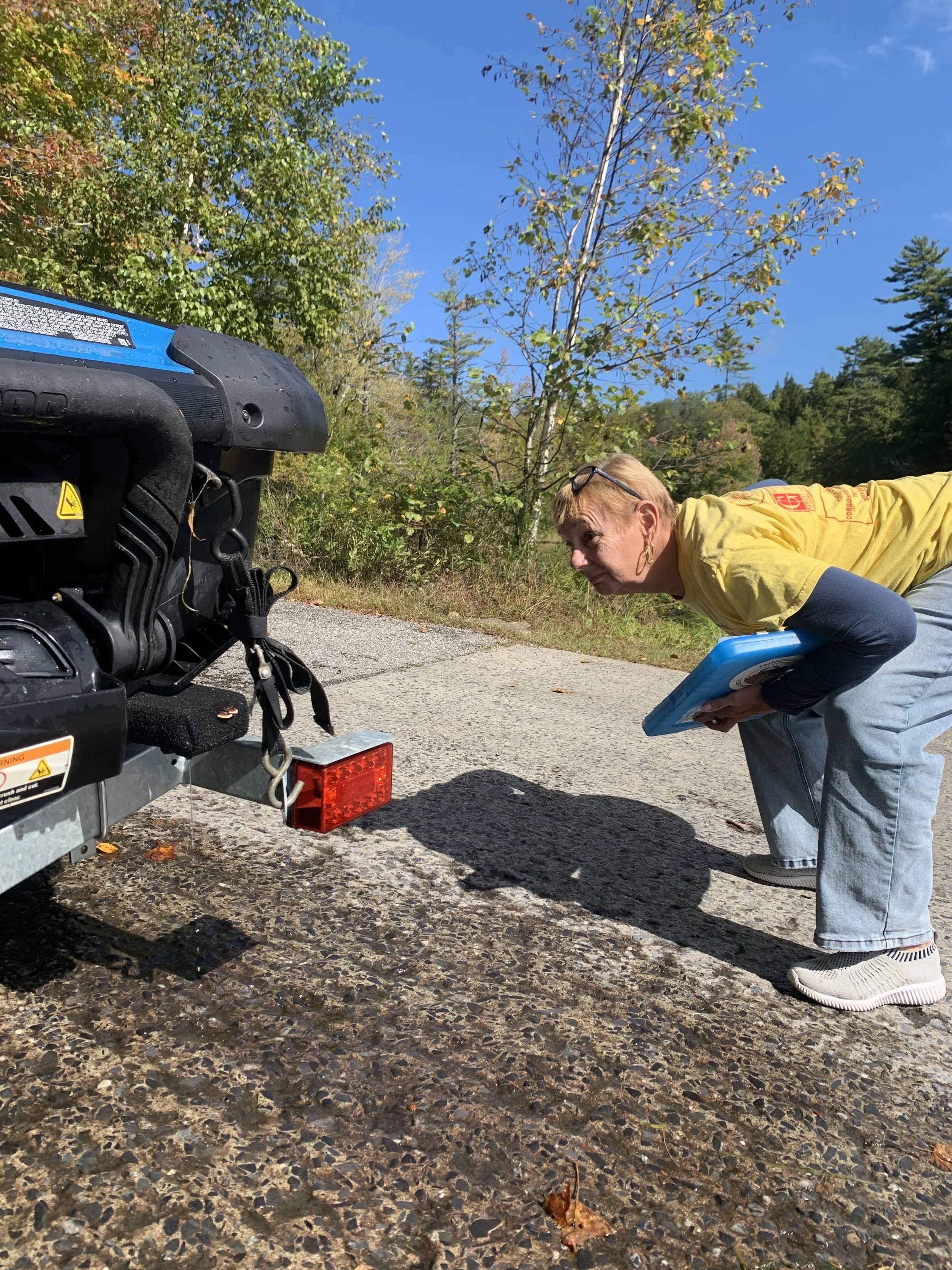 A woman wearing a yellow t-shirt and holding a blue clipboard crouched down to look under a boat that is on a trailer