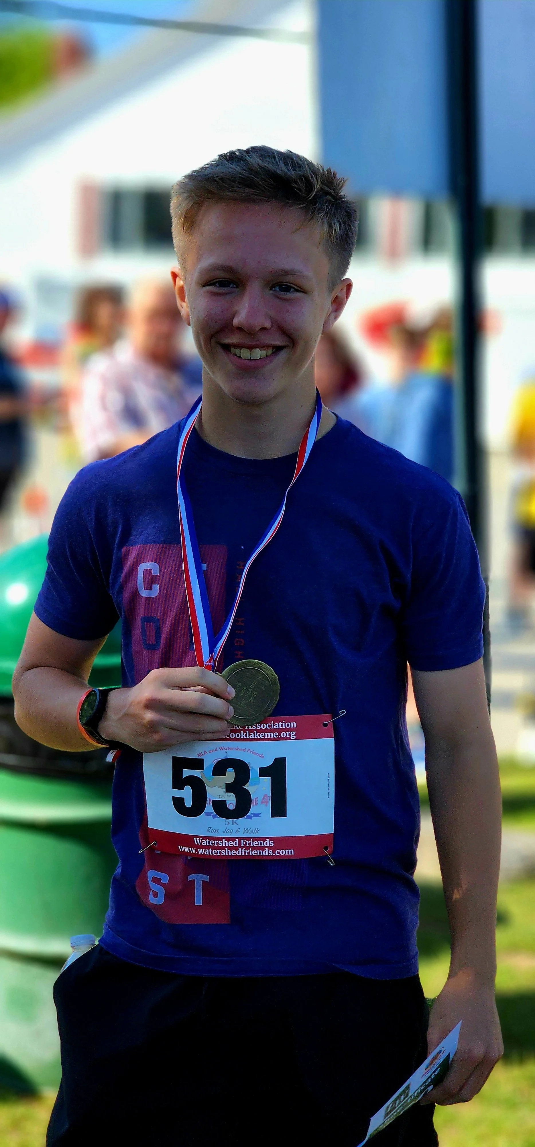 A man smiling at the camera, wearing an award medal, blue t-shirt, and black shorts