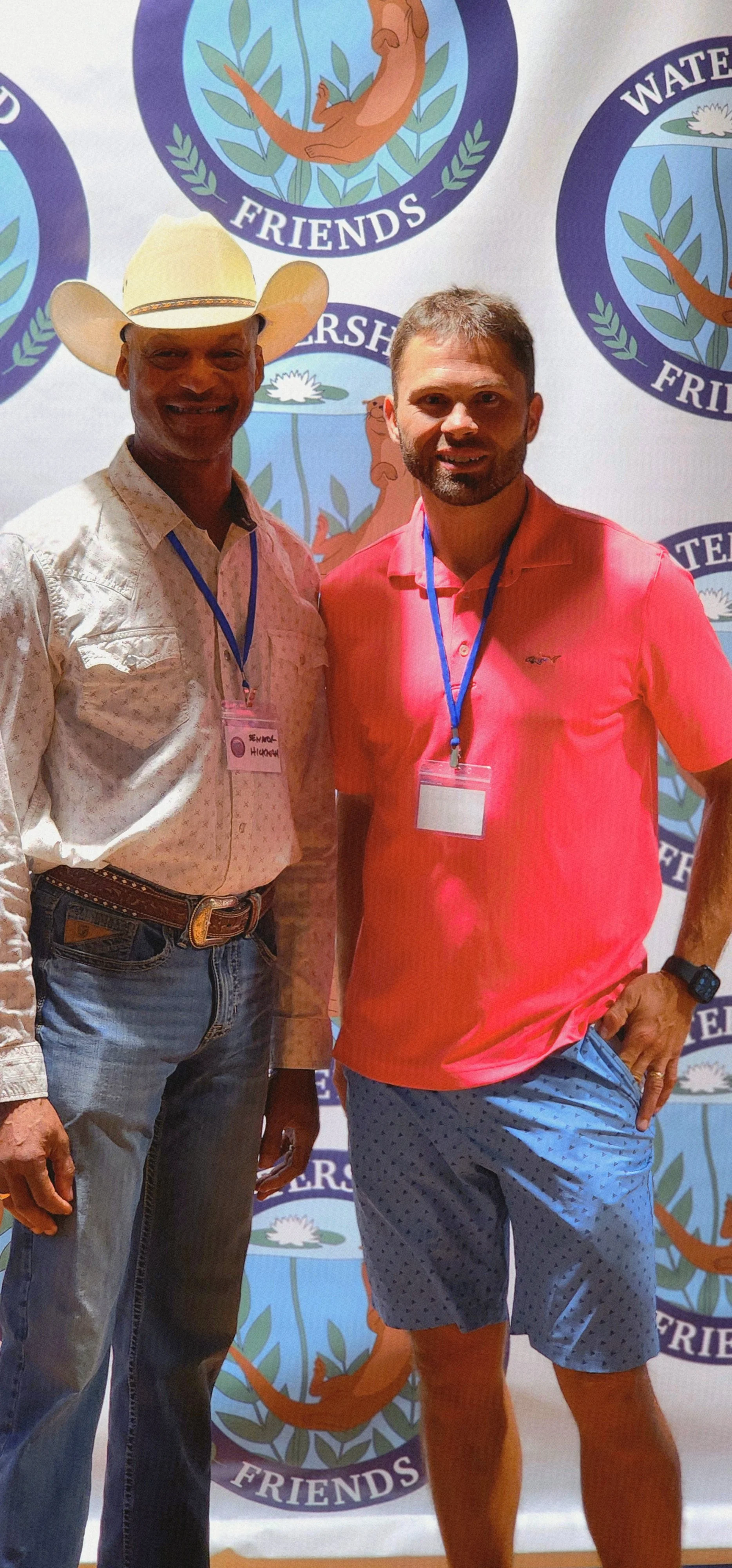 Two men, the one on the left wearing a white cowboy hat, white shirt, and jeans and the one on the right wearing a red polo shirt and blue shorts