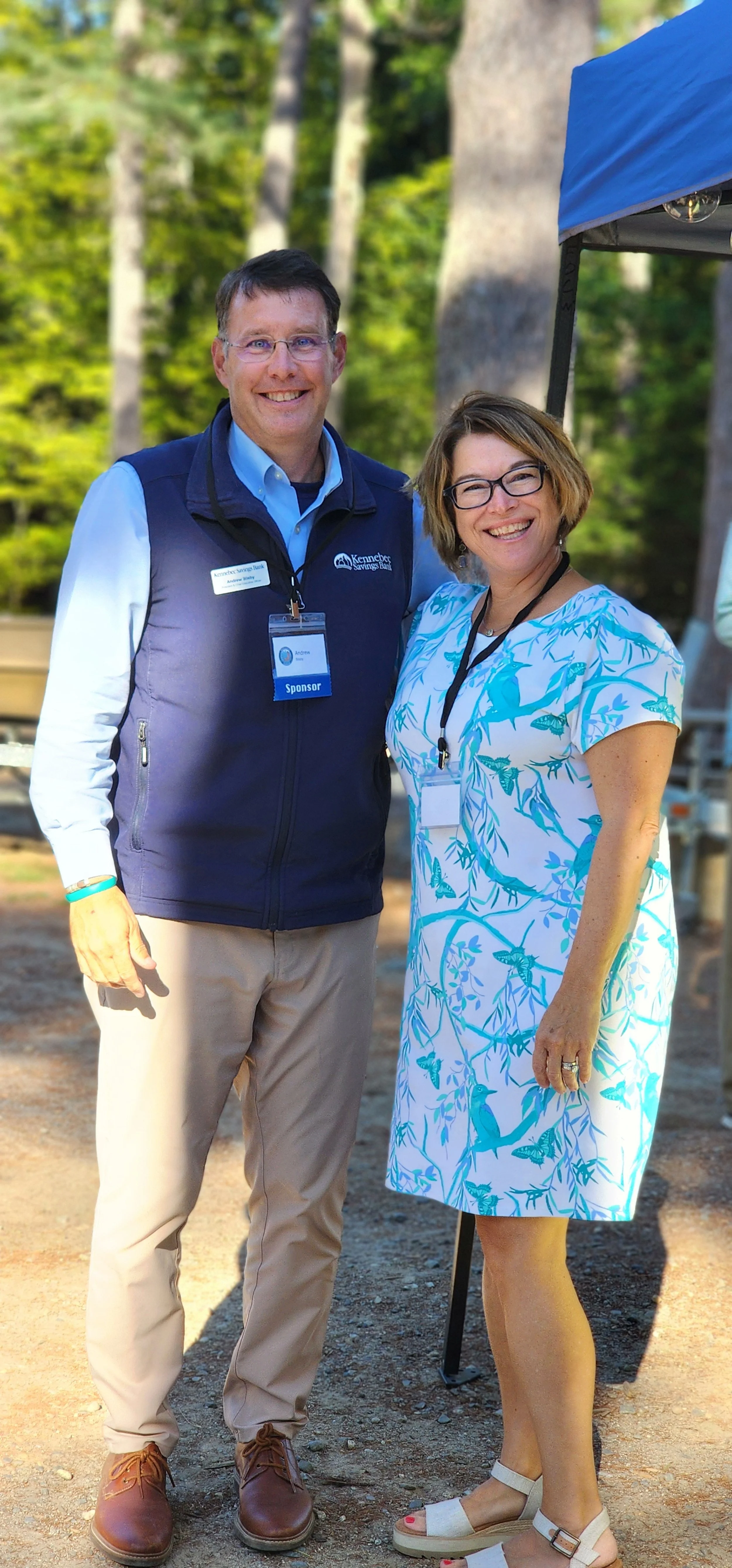 A man, wearing a dark blue vest over a light blue shirt and khaki shirts, and a woman, wearing a blue and white dress, smiling at the camera