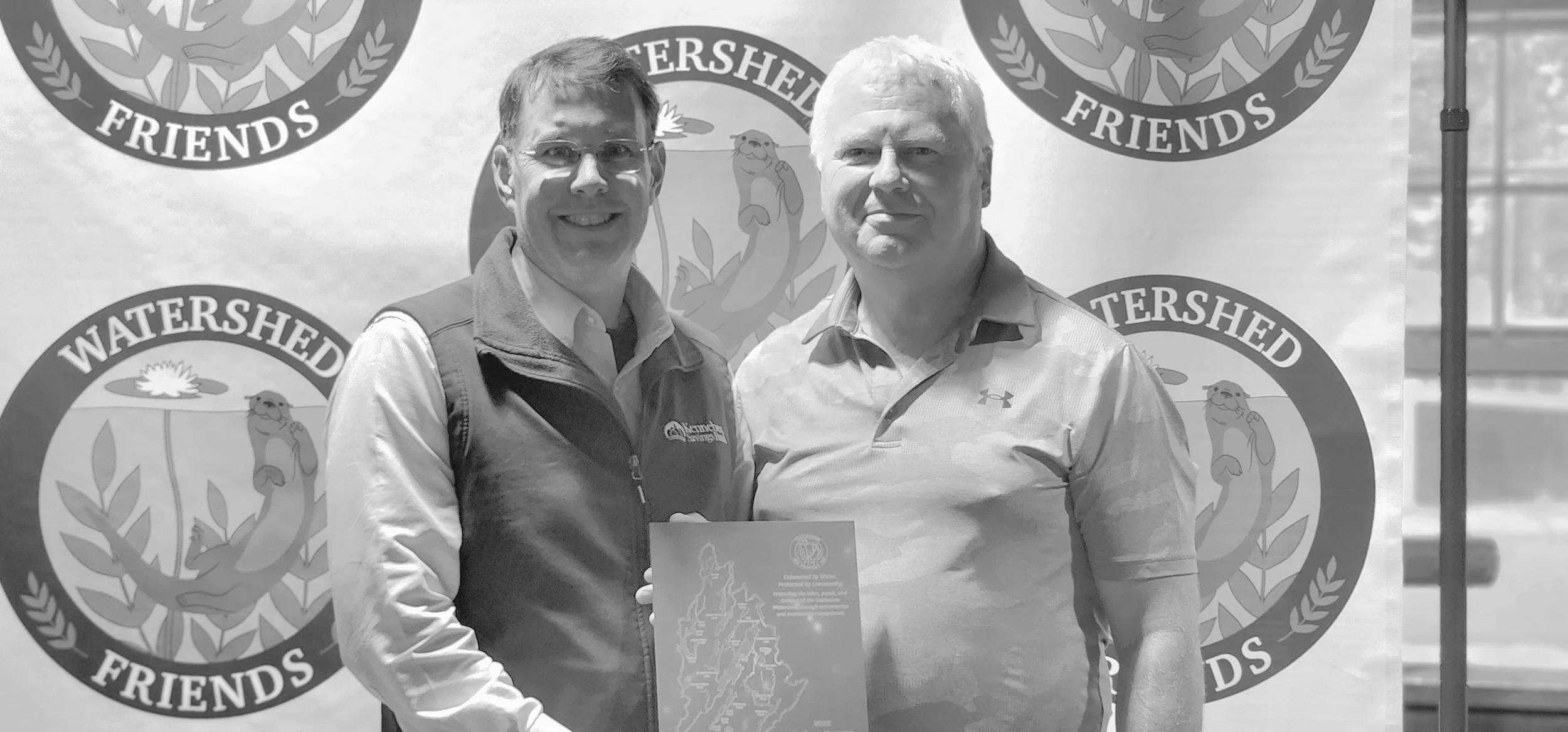 Two men, one wearing a vest over a long sleeve shirt and holding an award, the other wearing a polo shirt, smiling in front of a Watershed Friends logo backdrop