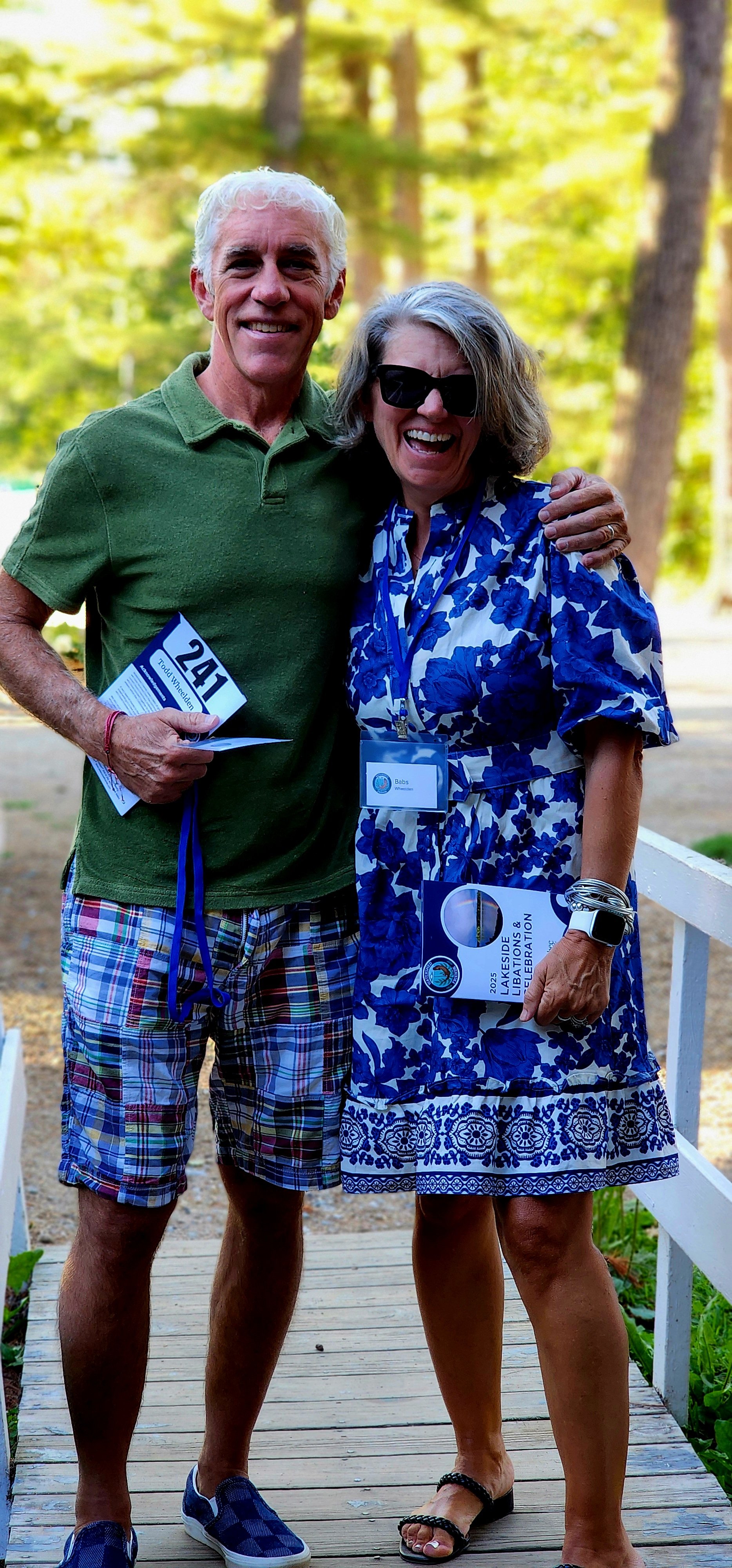A man, wearing a green polo and blue patterned shorts, and a woman, wearing a blue and white patterned dress, smiling at the camera