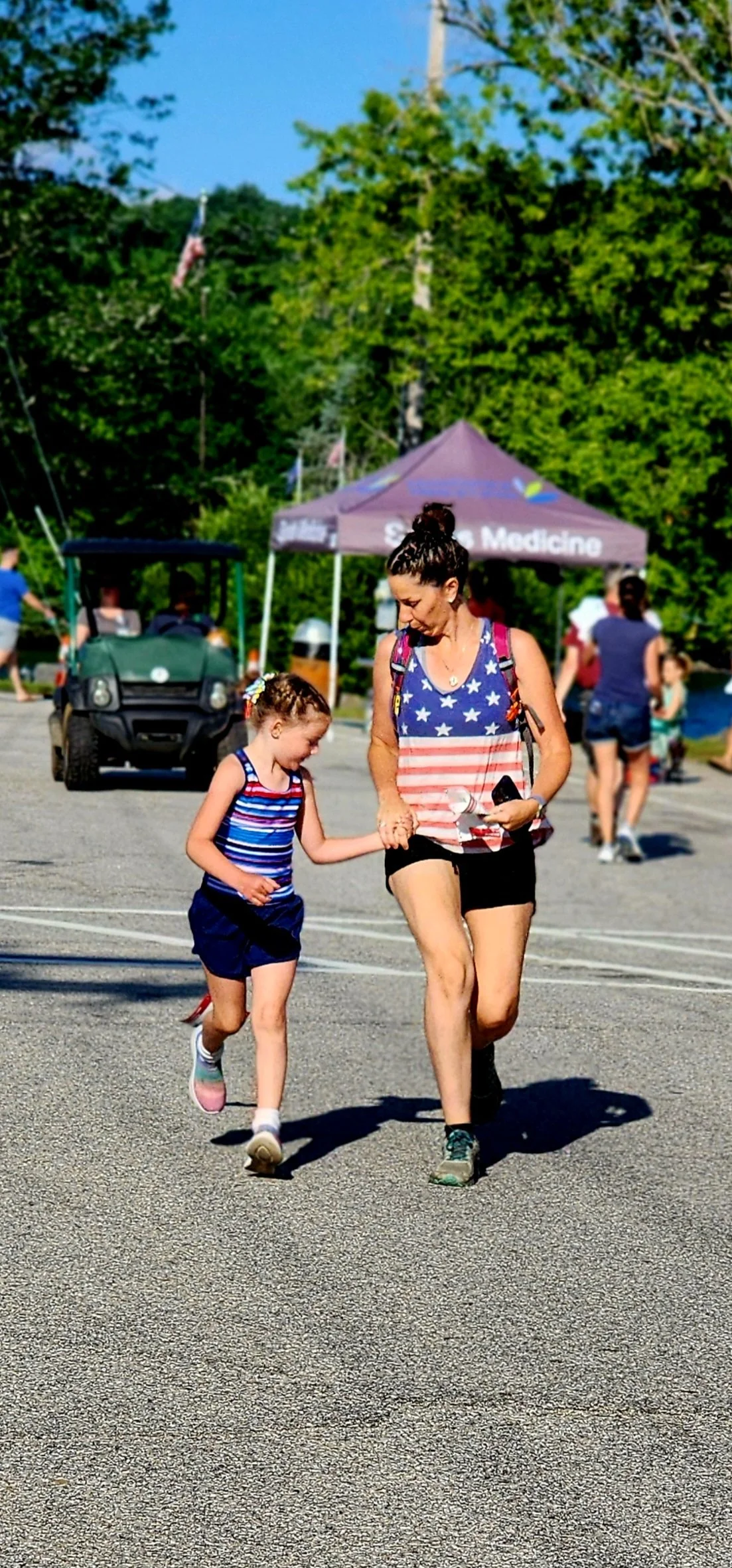 A girl, wearing a striped tank top and black shorts, holding hands a jogging with a woman wearing an Amercan flag tank top and black shorts
