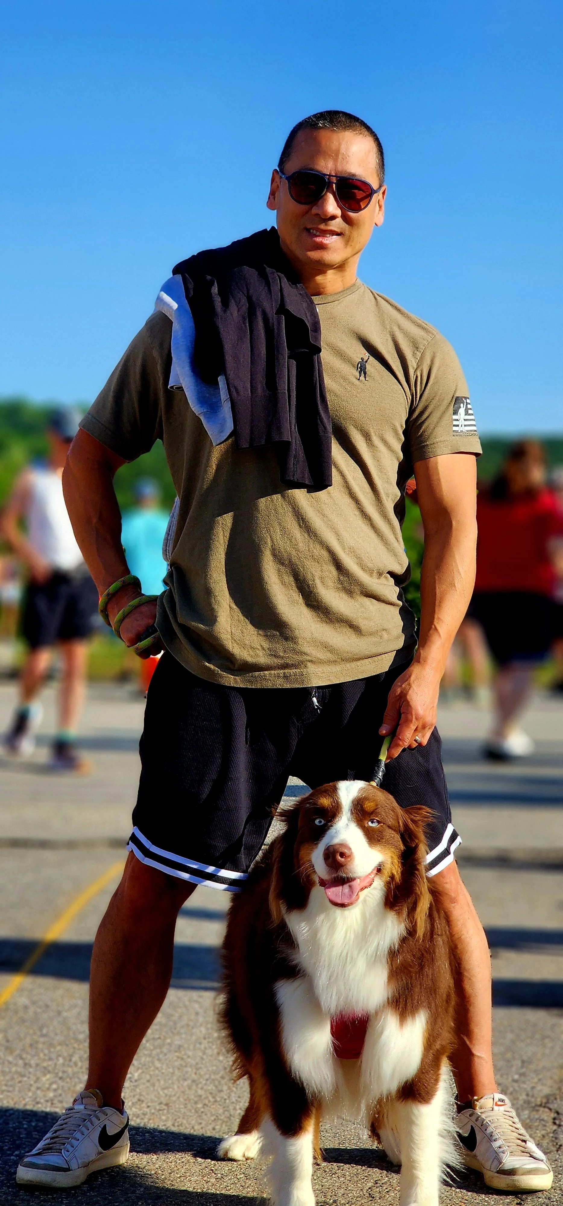A man, wearing a beige shirt and black shorts, and a dog smiling at the camera