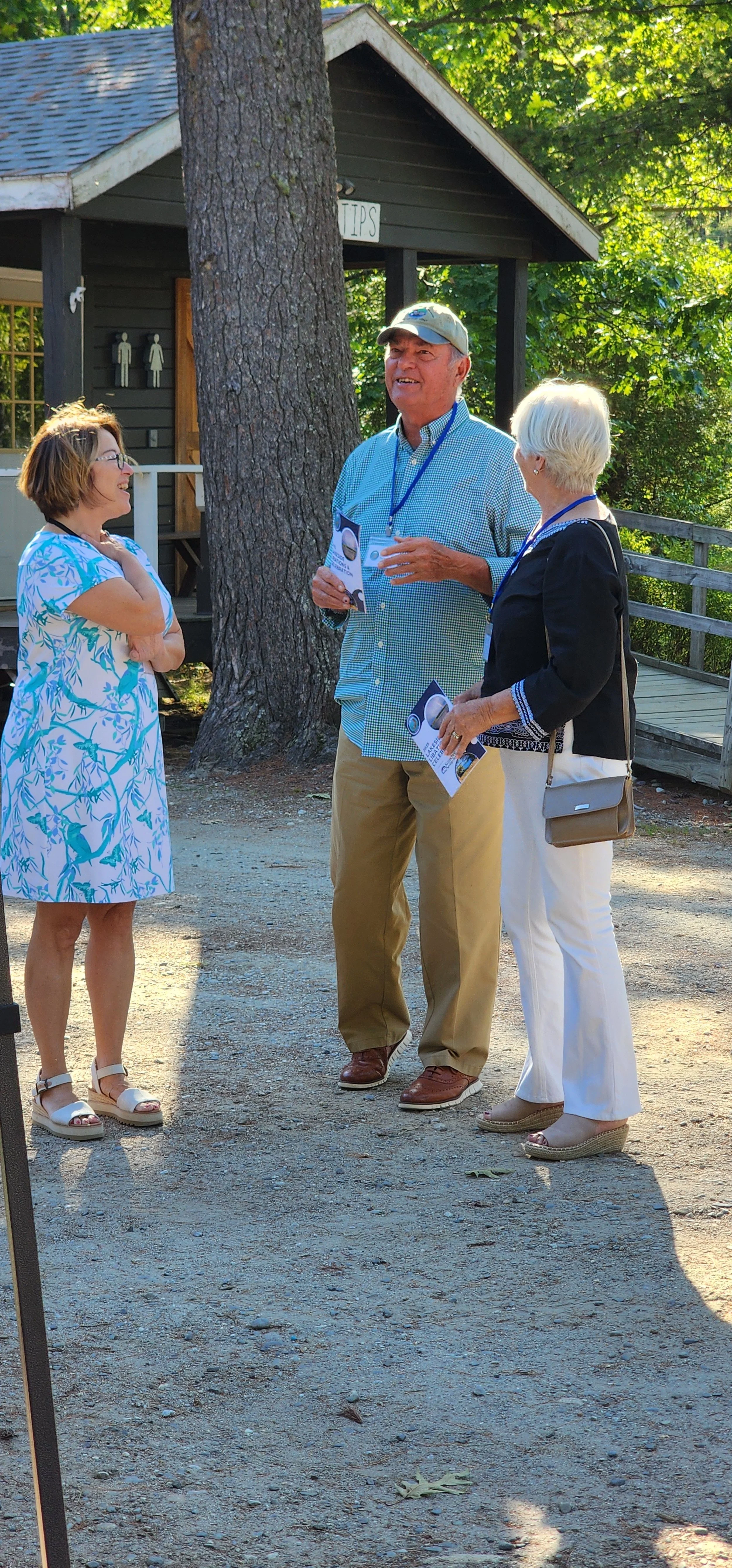 Three people, a woman in a blue and white dress on the left, a man in a blue long sleeve shirt and khaki pants in the middle and a woman wearing a black long sleeve shirt and white pants, chatting