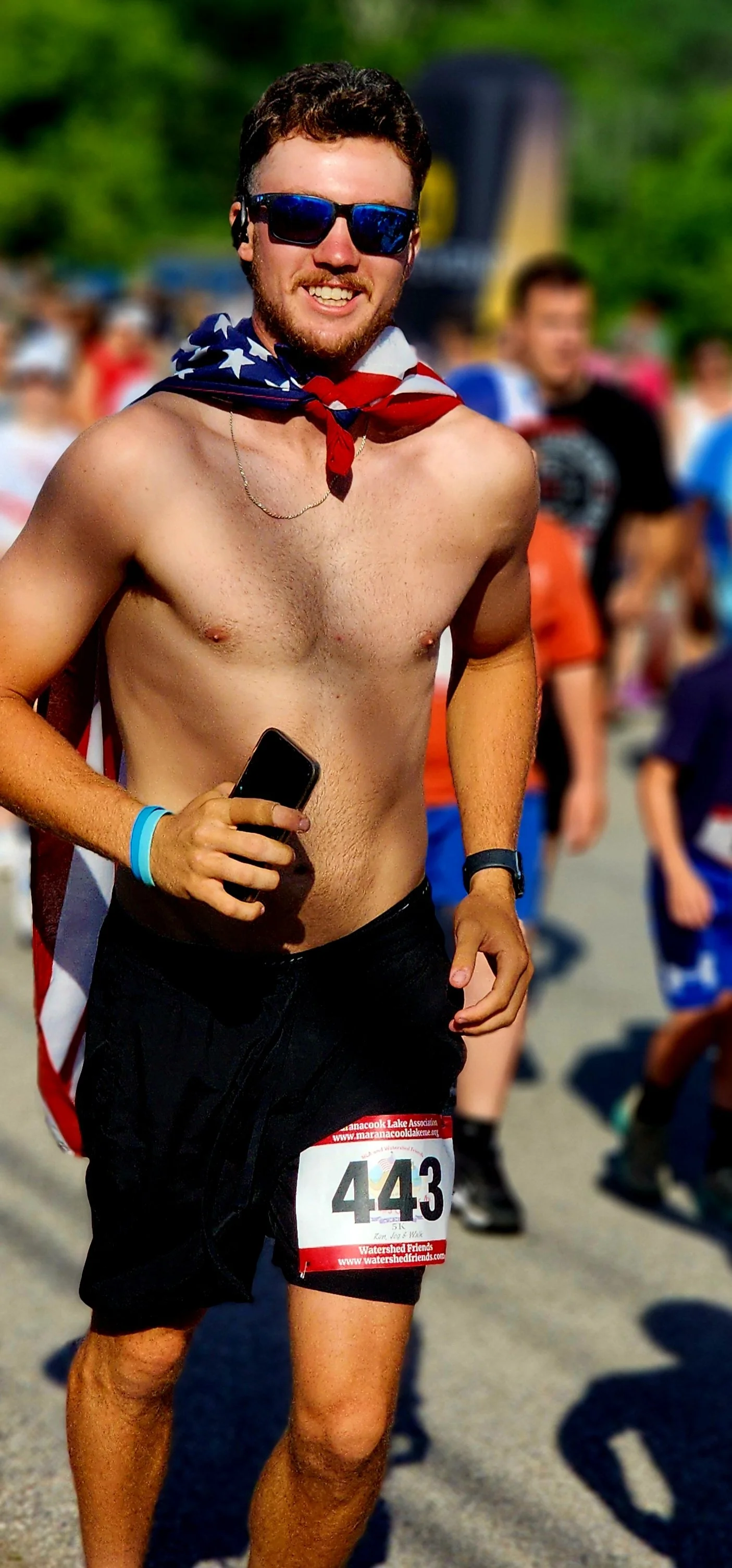 A shirtless man wearing an American flag as a cape and back shorts, jogging and smiling at the camera
